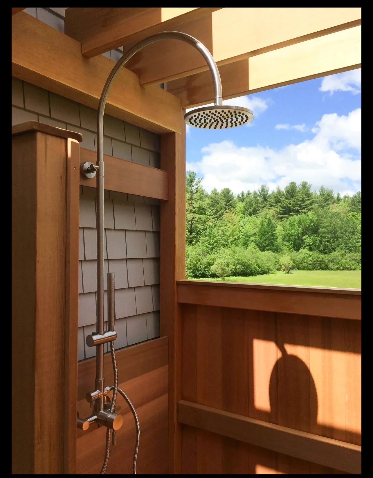 An outdoor shower with cedar construction and a picturesque window-view of mowed grass field and a lush green tree line.