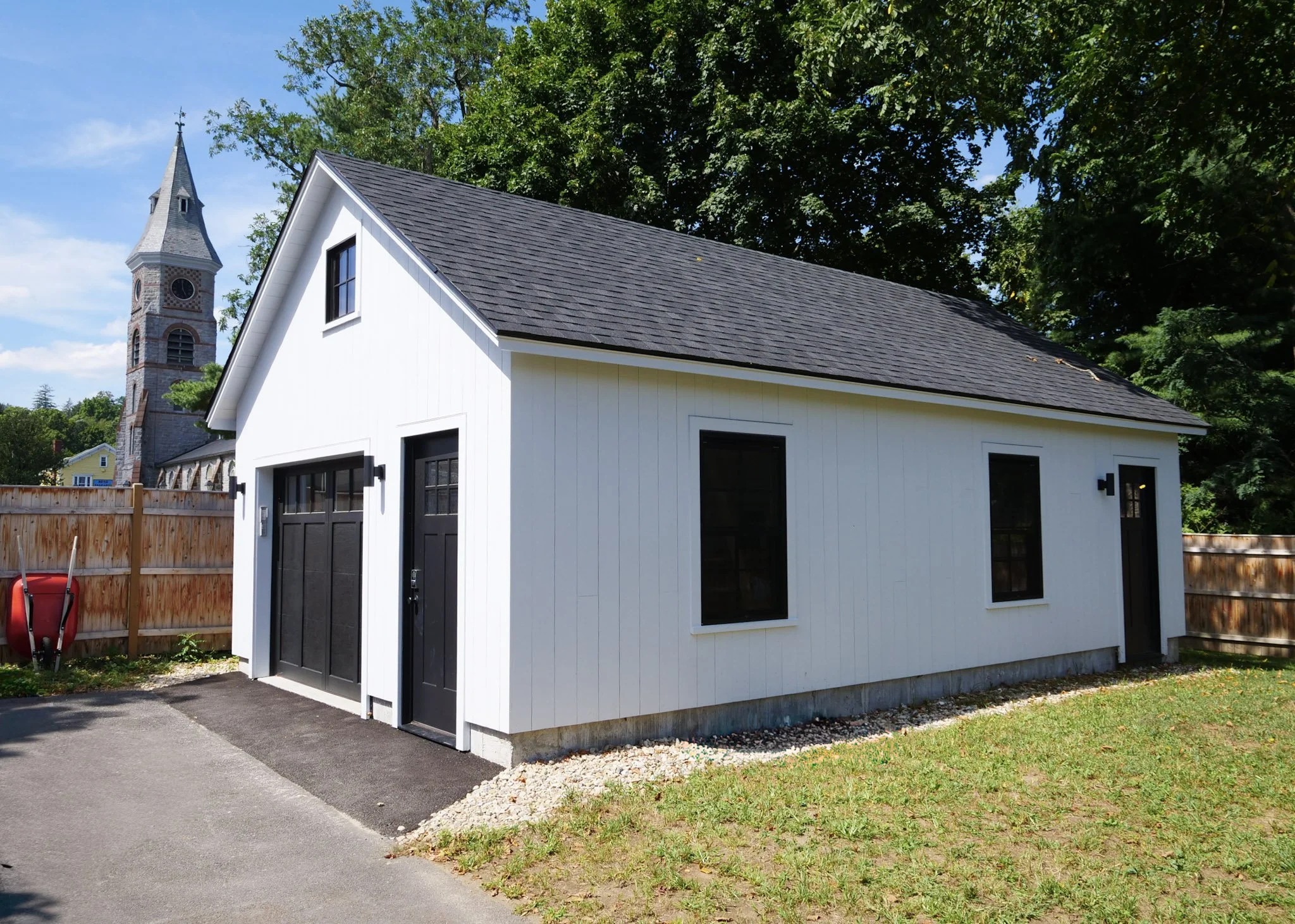 A small white garage with black doors and windows, a sloped roof, surrounded by a wooden fence, with a church steeple visible in the background, and green trees under a blue sky.