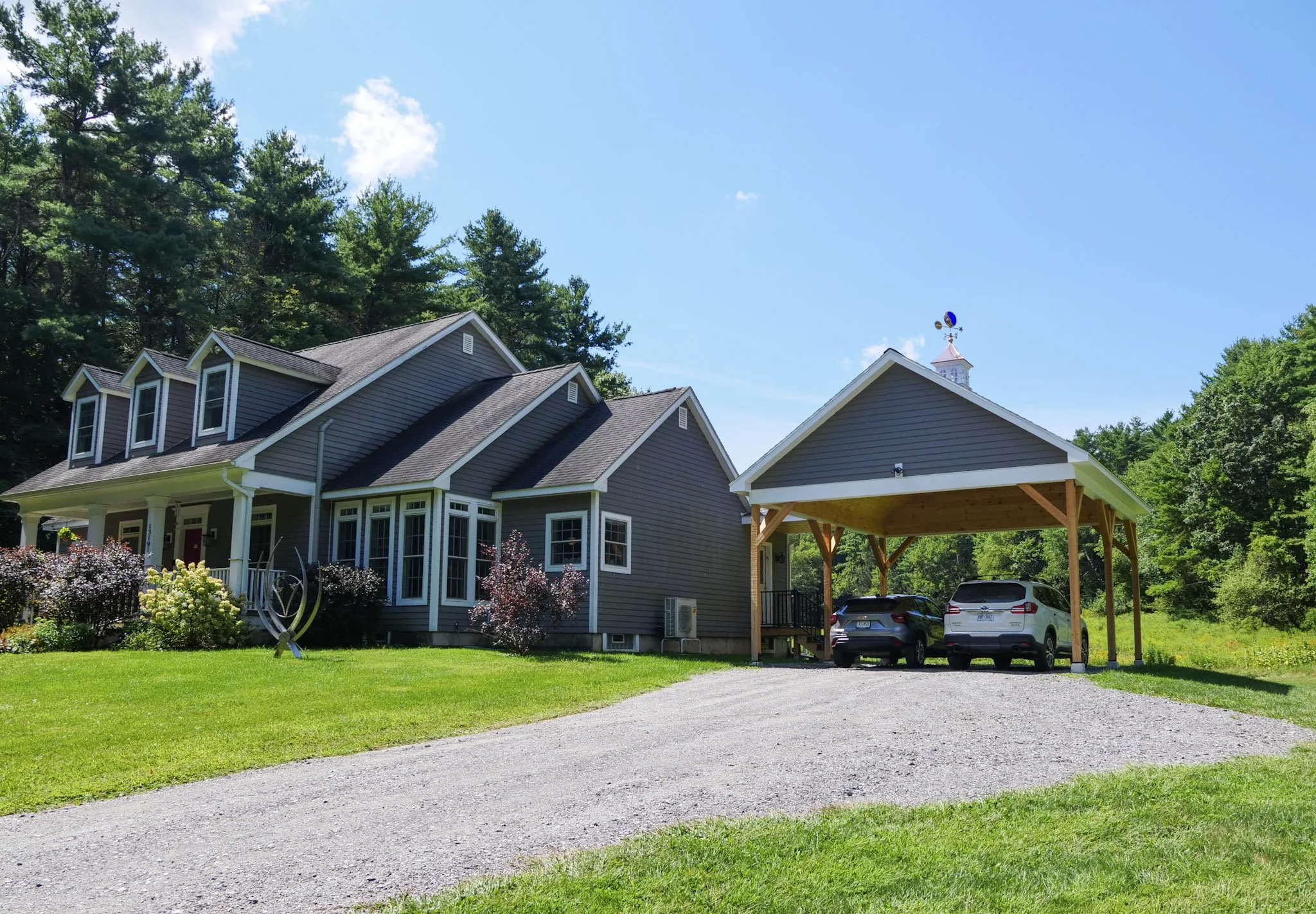 A house with gray siding and white trim, surrounded by a green lawn and colorful plants, with a gravel driveway leading to a parking area with two cars and a wooden carport, and tall trees in the background under a bright blue sky with a few clouds.