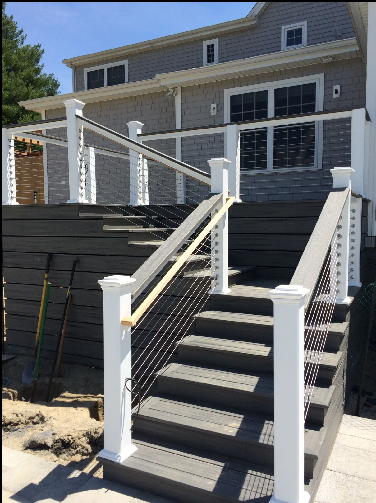 Newly constructed wooden staircase with white railings and cable spindles leading up to a second-story deck of a gray house with sliding glass doors and windows.