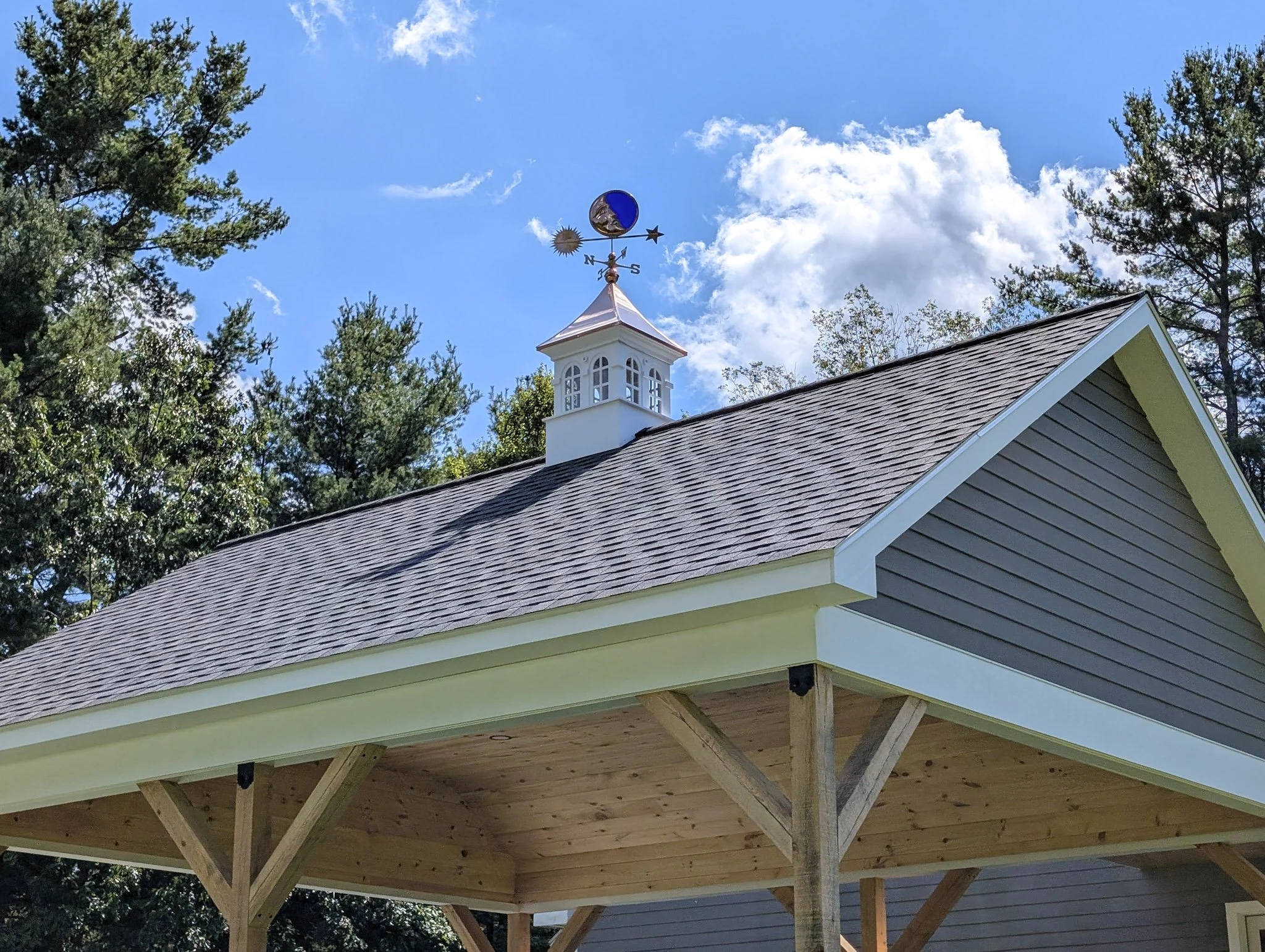 A building with a pitched roof under construction, with a weather vane on top showing moon, sun, stars, and cardinal directions, surrounded by trees and a partly cloudy sky.