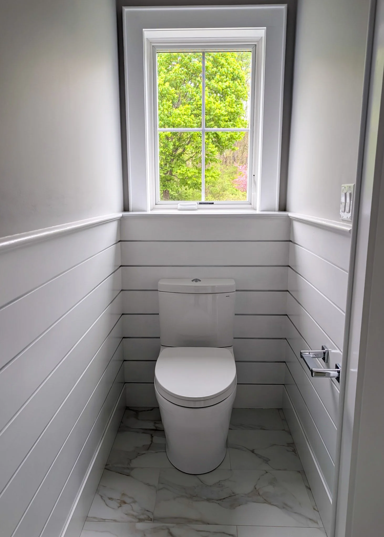 Toilet in water closet against white wainscotting and light grey walls, sitting on white marble floor with dramatic grey veins