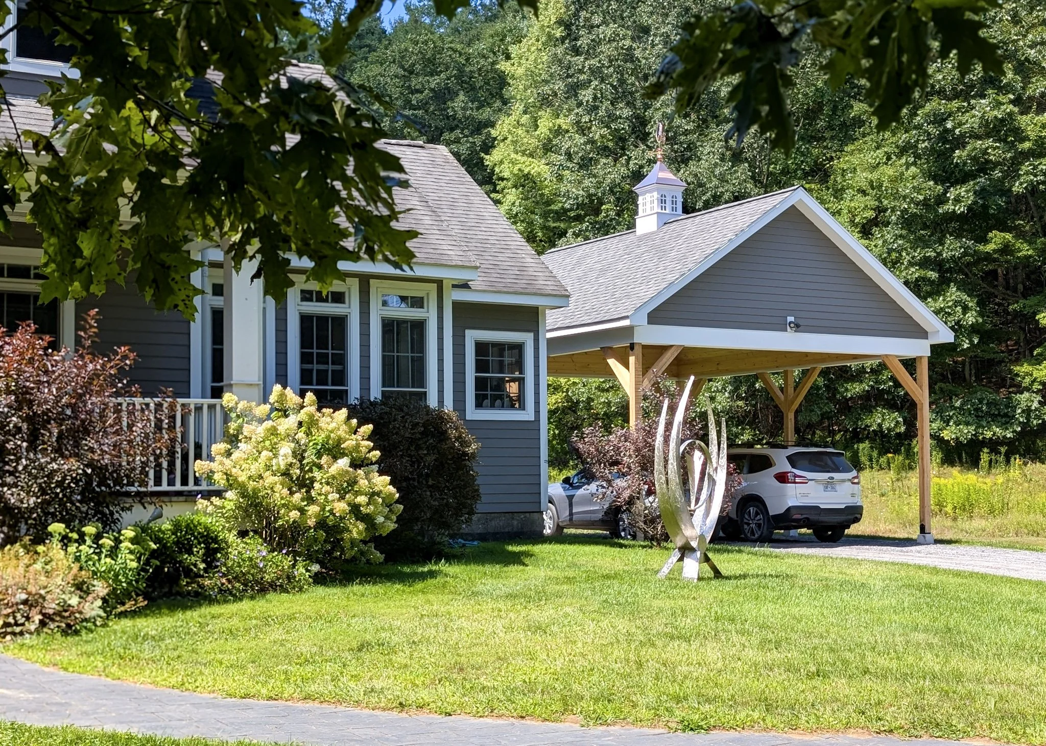 Front yard of a house with lush green lawn, colorful bushes, trees, a sculpture, and a car parked near a garage under a wooden structure.
