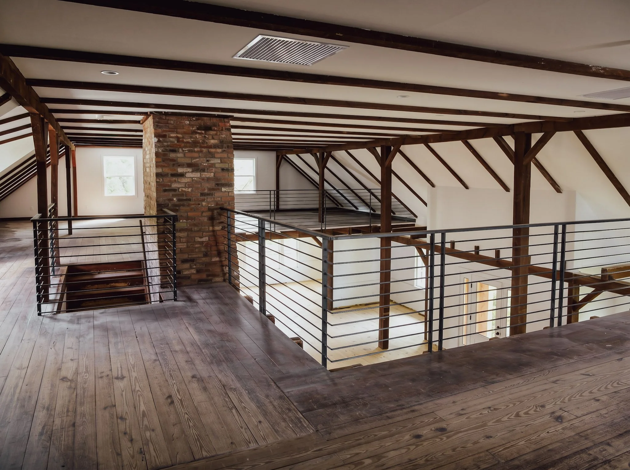Open loft area with wooden floors, exposed wooden beams on the ceiling, brick pillar, large windows, and metal railing around the edge.