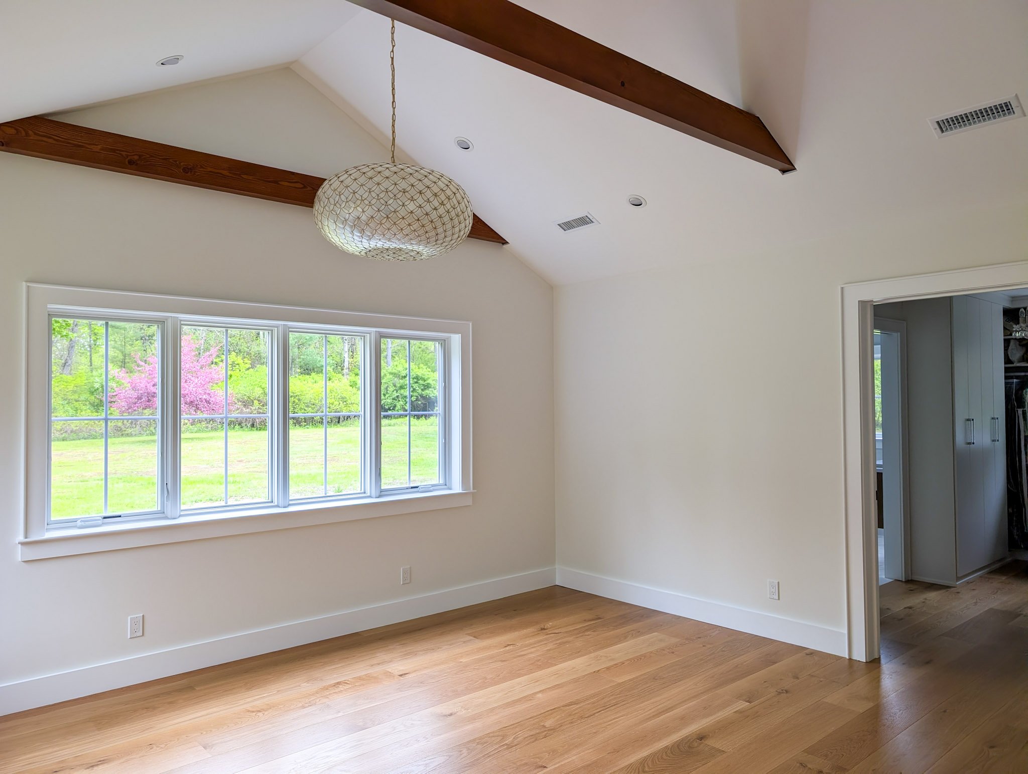 Empty room with large window overlooking a green backyard, wooden floor, white walls, and a ceiling light fixture with a decorative cover.
