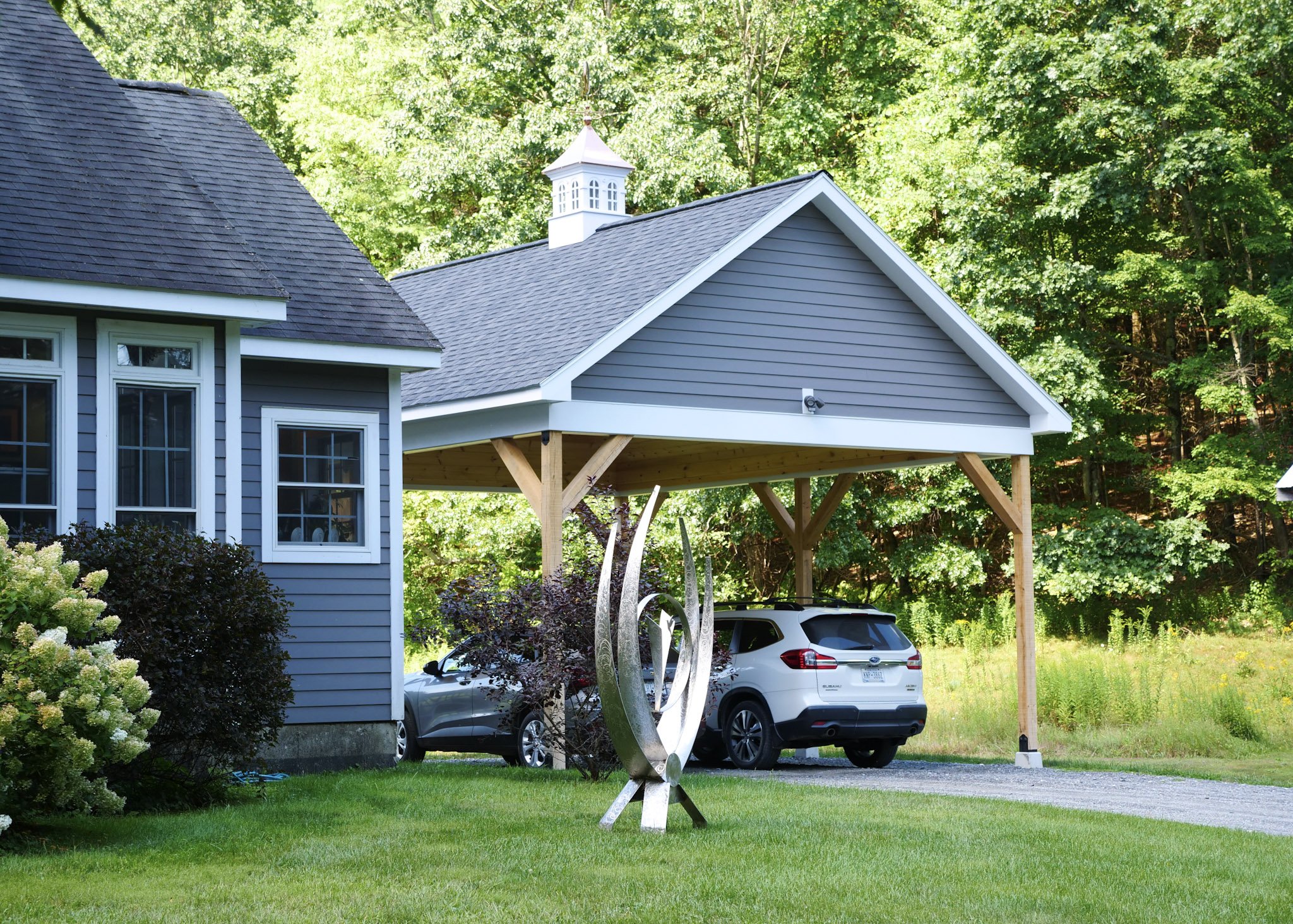 A blue-gray house with white trim and large windows, a gravel driveway with two parked cars, a grass lawn, trees in the background, and a small white cupola on the roof.
