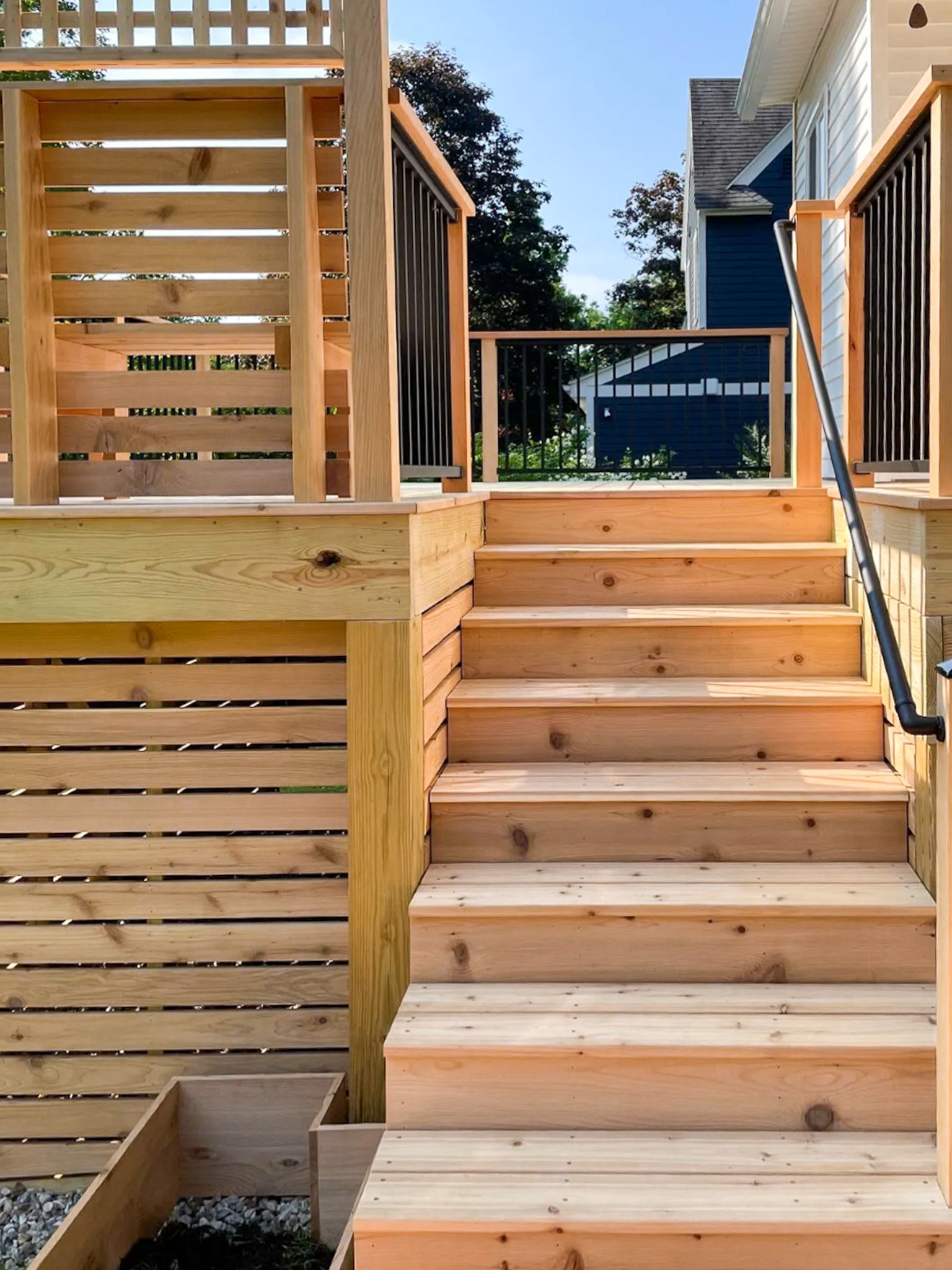 Newly built wooden stairs leading up to a deck with a modern railing, in a backyard with trees and neighboring houses visible in the background.
