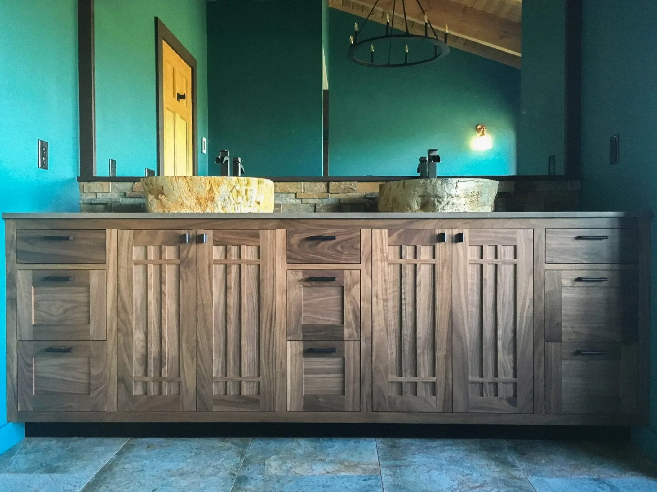 View of a bathroom vanity with two stone sinks, wooden cabinetry, and teal-colored walls, with a chandelier hanging from the ceiling and a wall-mounted light fixture.