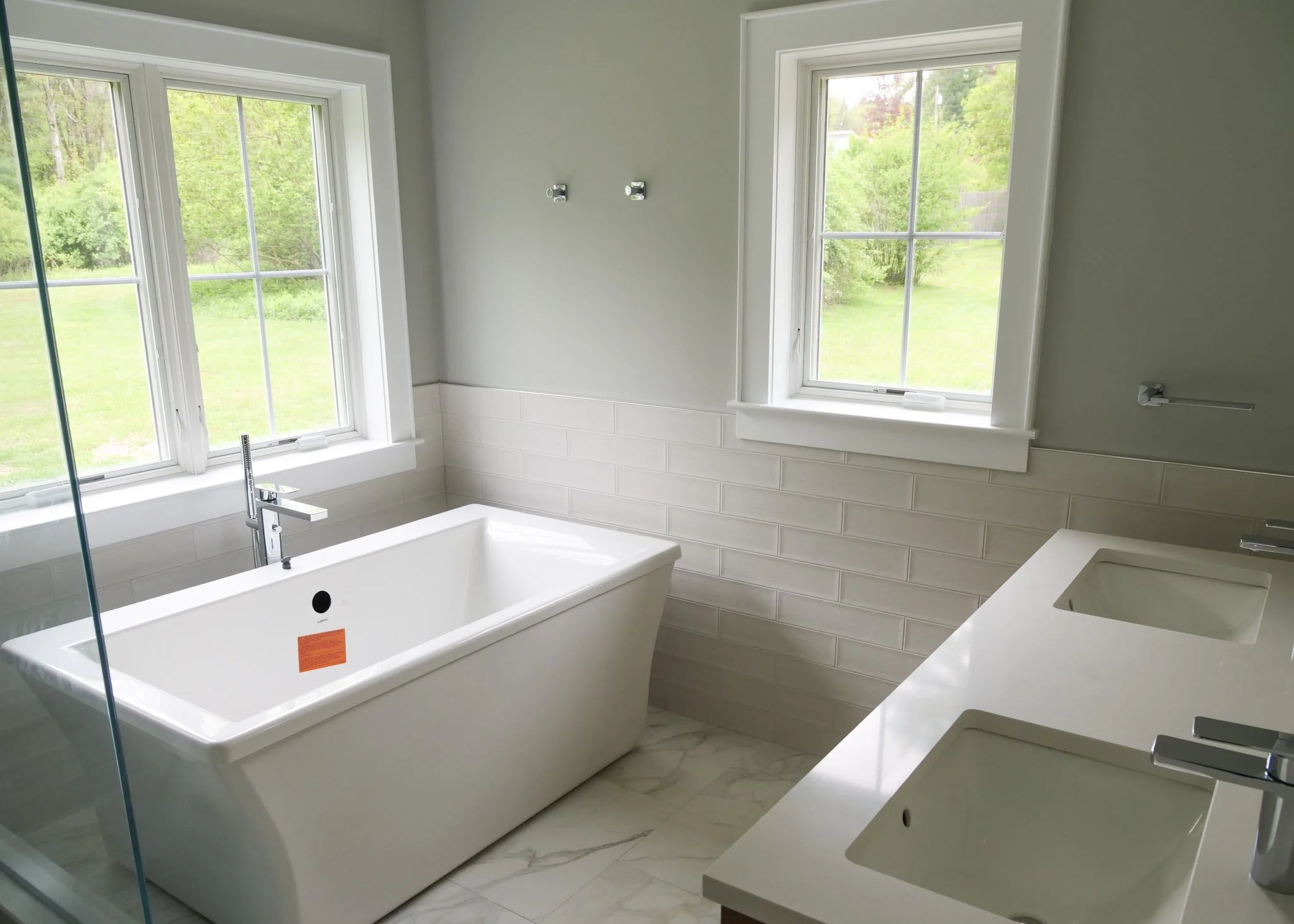 A bathroom with two large windows, a bathtub, a sink with a faucet, and a countertop with multiple sinks, all with modern fixtures. Outdoor greenery is visible through the windows.