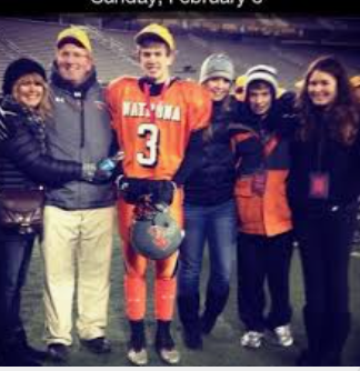 Group of six people posing together outdoors, one in football uniform with a helmet and ball, five in casual jackets and hats.