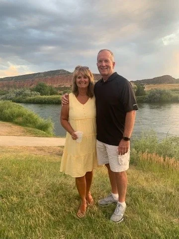 A smiling man and woman standing together outdoors by a river with a grassy area and hills in the background. The woman is holding a cup, and the man has his arm around her.