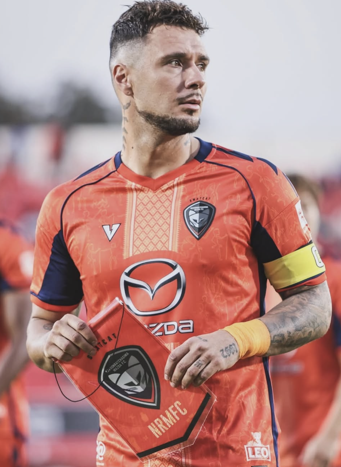 A male soccer player holding a team flag, wearing an orange and black uniform with sponsorship logos and a captain's armband, standing outdoors during a game.