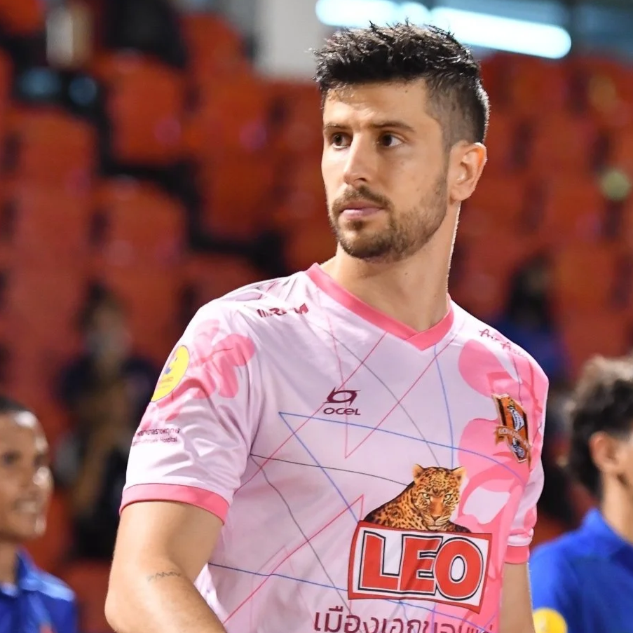A man with dark, spiky hair and facial hair wearing a pink soccer jersey with leopard and LEO logos stands on a soccer field with blurred spectators in the background.