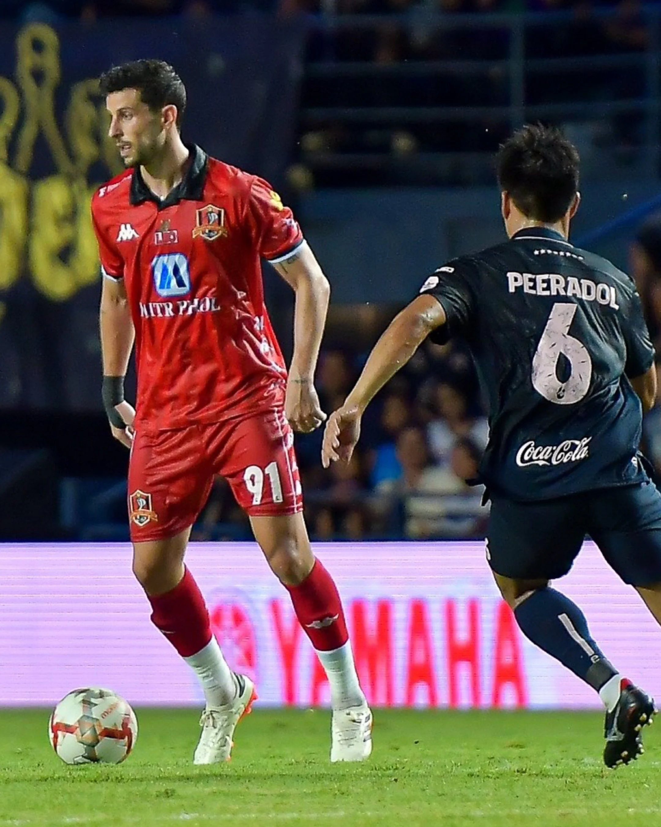 Two soccer players holding hands on the field. The player on the left is wearing a red uniform with the number 21. The player on the right has "PEERADOL" and the number 6 on the back of his black uniform. A soccer ball is on the ground near their feet.