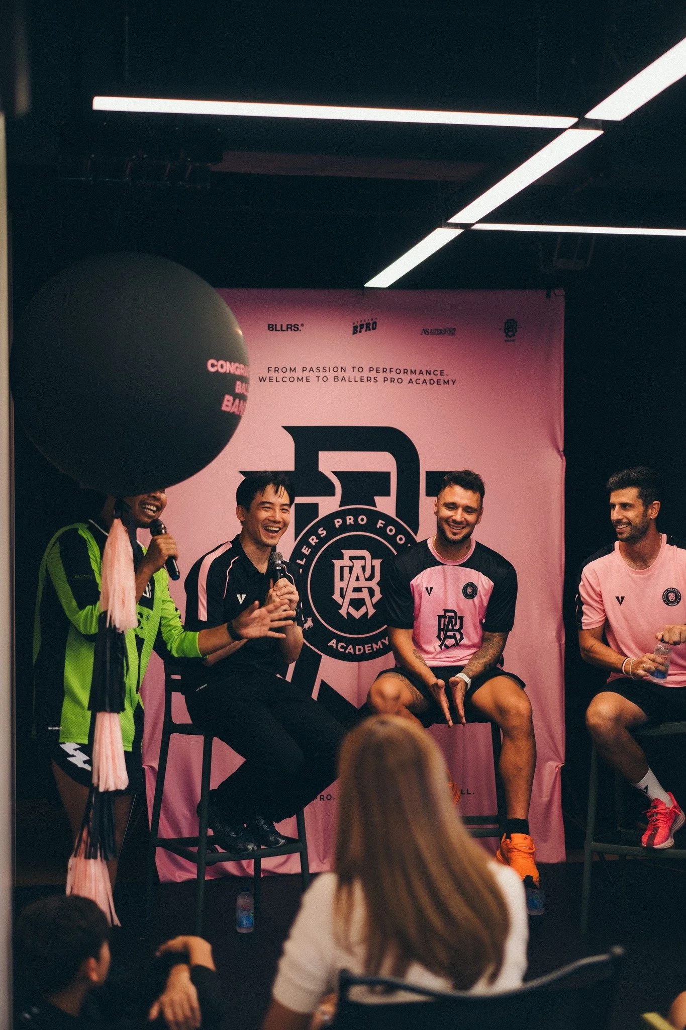 A group of men sitting on a stage, smiling and talking. They are wearing sports or team jerseys and are participating in an event related to Ballers Pro Academy. One man is holding a microphone, and there is a pink banner in the background with logos and text.