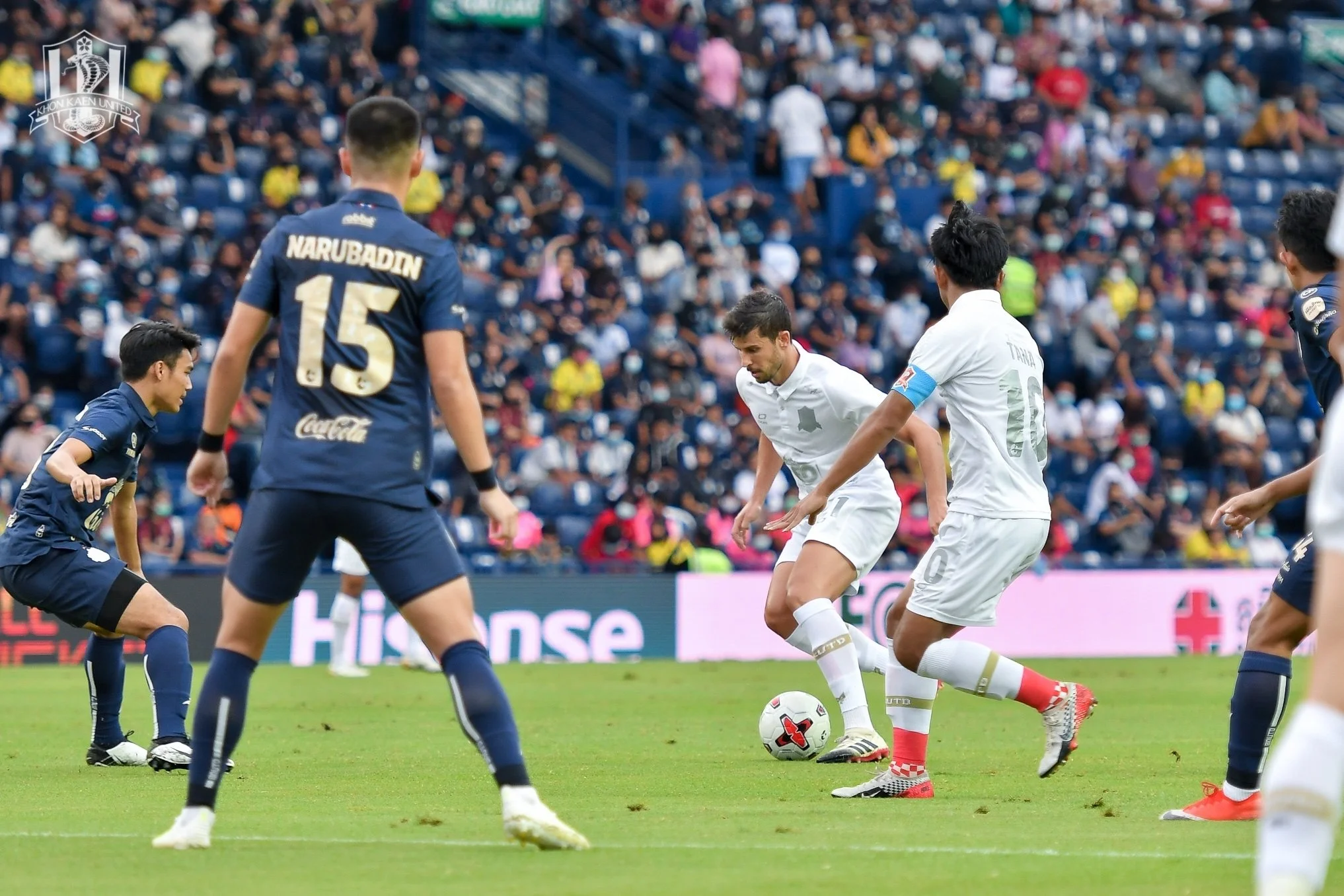 Soccer players on the field during a game with a crowd in the stands. A player in white is controlling the ball while players in blue are nearby.