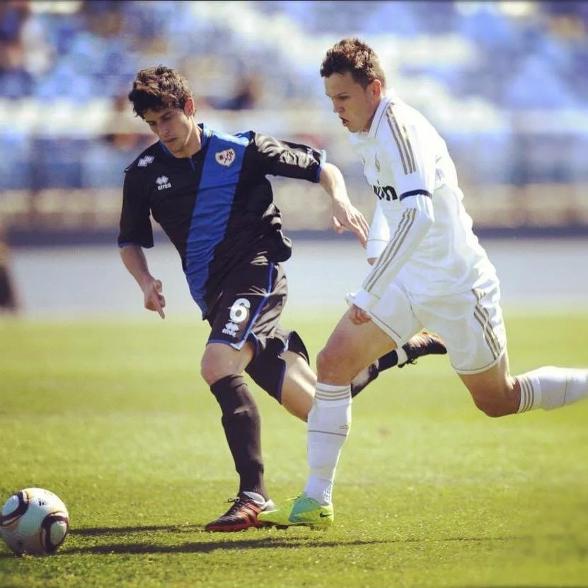 Two young male soccer players compete for possession of the ball on a soccer field, one wearing a black and blue jersey, the other in a white jersey.