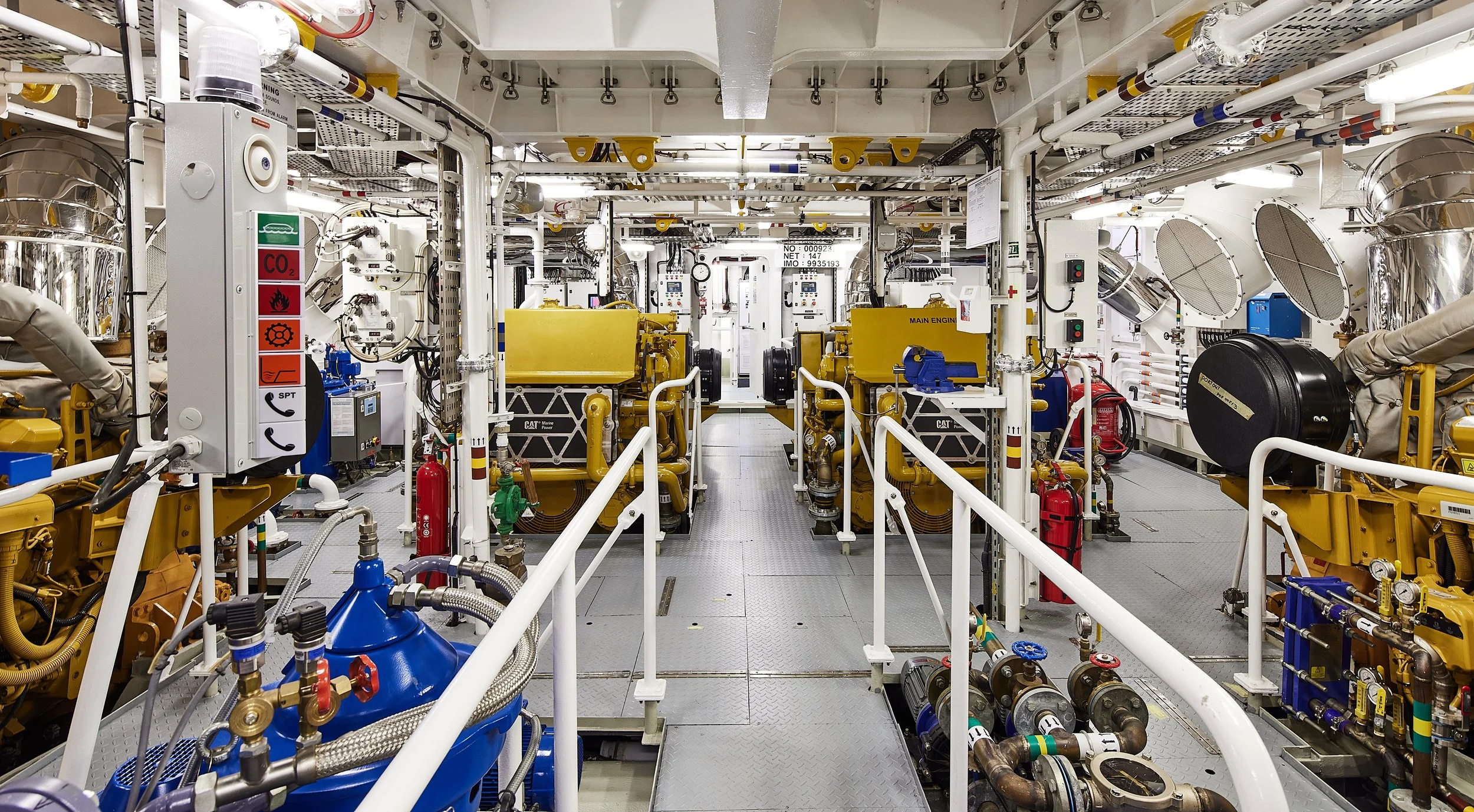 Interior of an industrial machine room with yellow engines, blue and red pipes, gauges, and safety equipment.