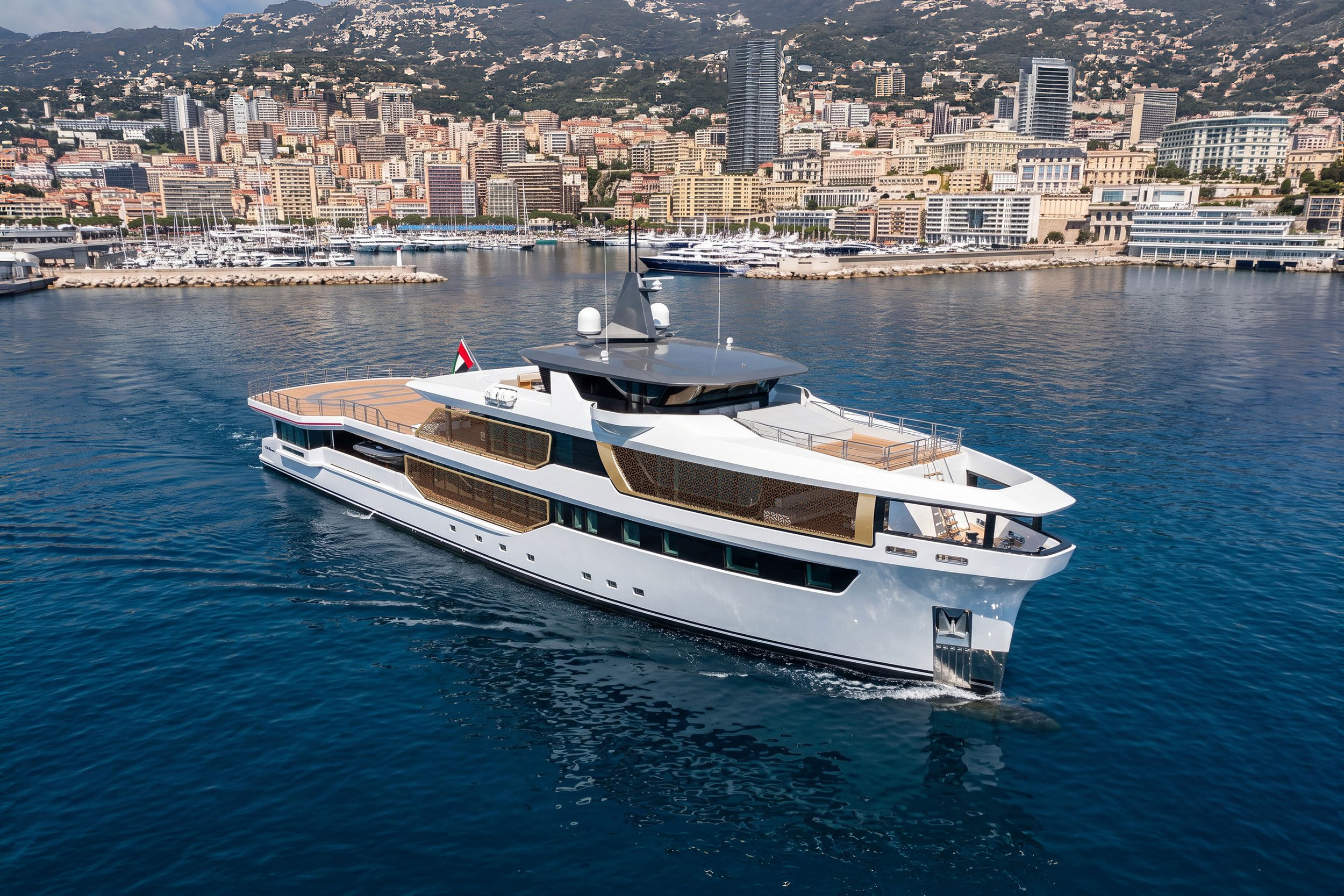 Luxury yacht with a white hull and wooden deck sailing in a harbor, with a cityscape and mountains in the background.