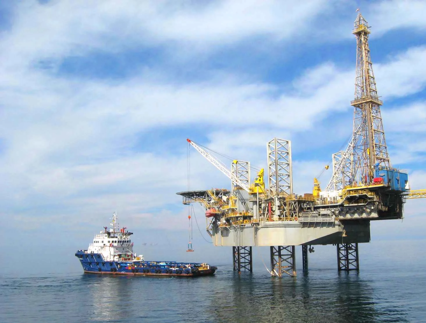 Offshore oil rig with a tugboat nearby in calm water under a partly cloudy sky.