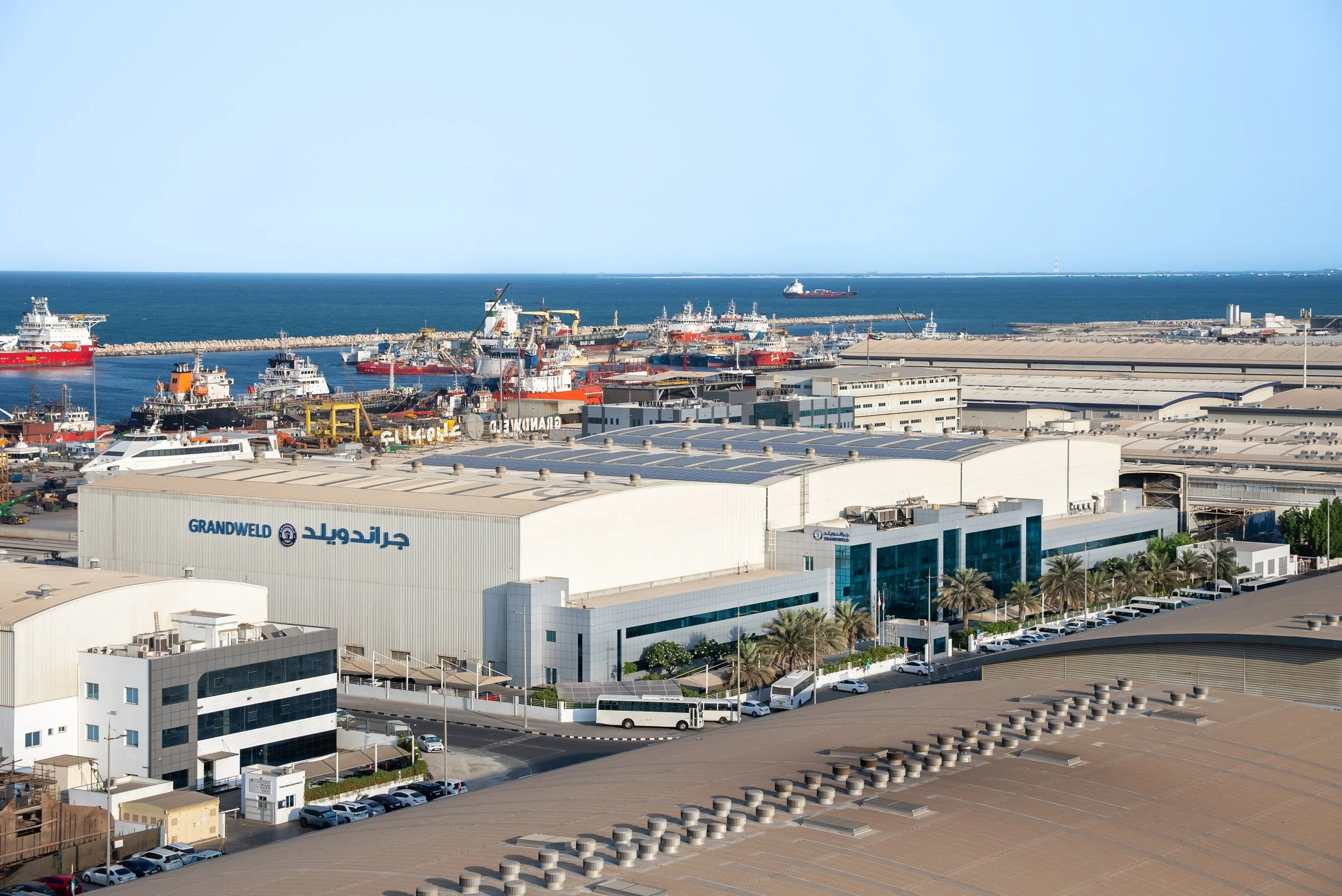 A port with multiple ships docked, industrial buildings in the foreground with a logo that reads 'Grandweld' in English and Arabic, and a clear blue sea with a cargo ship in the distance.