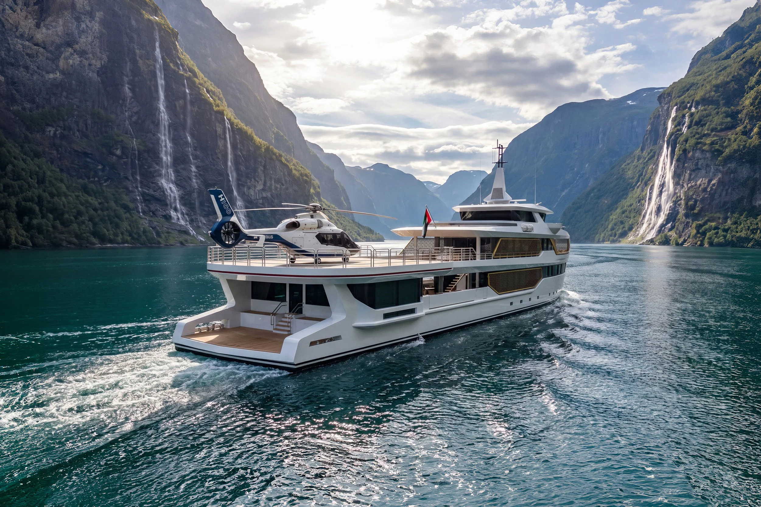 A luxurious white yacht with a helicopter on its deck sailing through a fjord with steep, green, rocky cliffs and waterfalls on both sides. The sky is partly cloudy and the water is calm.