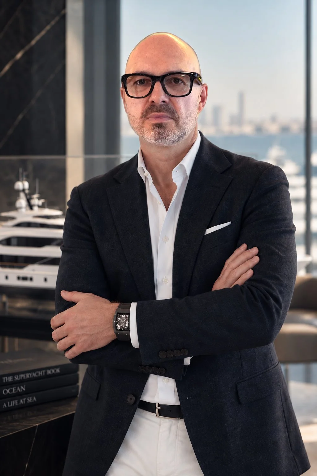 Bald man with glasses in a business suit standing with arms crossed in office with a view of the skyline and yachts outside.