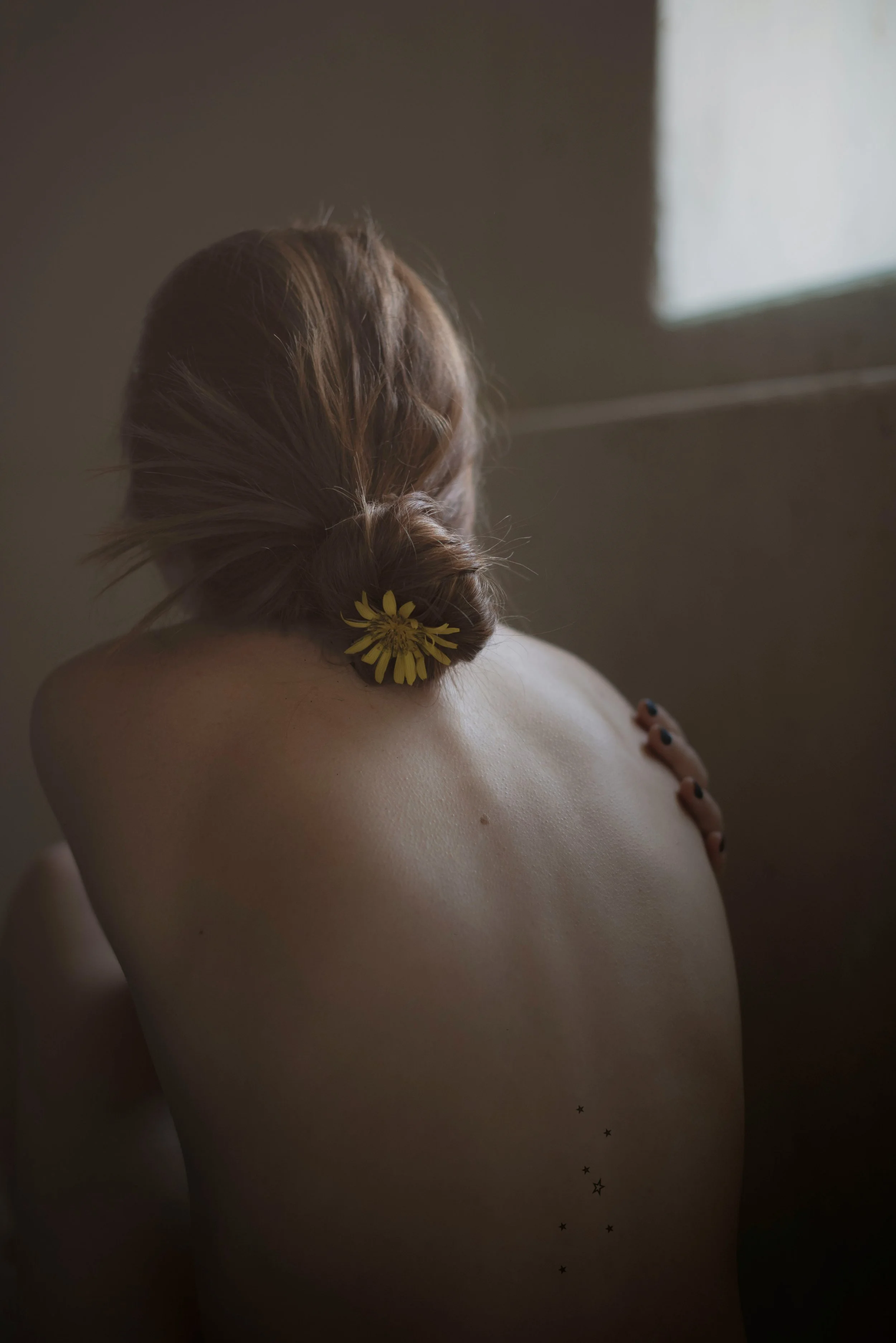 Back view of a woman with red hair styled in a low bun, wearing a yellow flower in her hair, sitting with her back to the camera in a softly lit room.