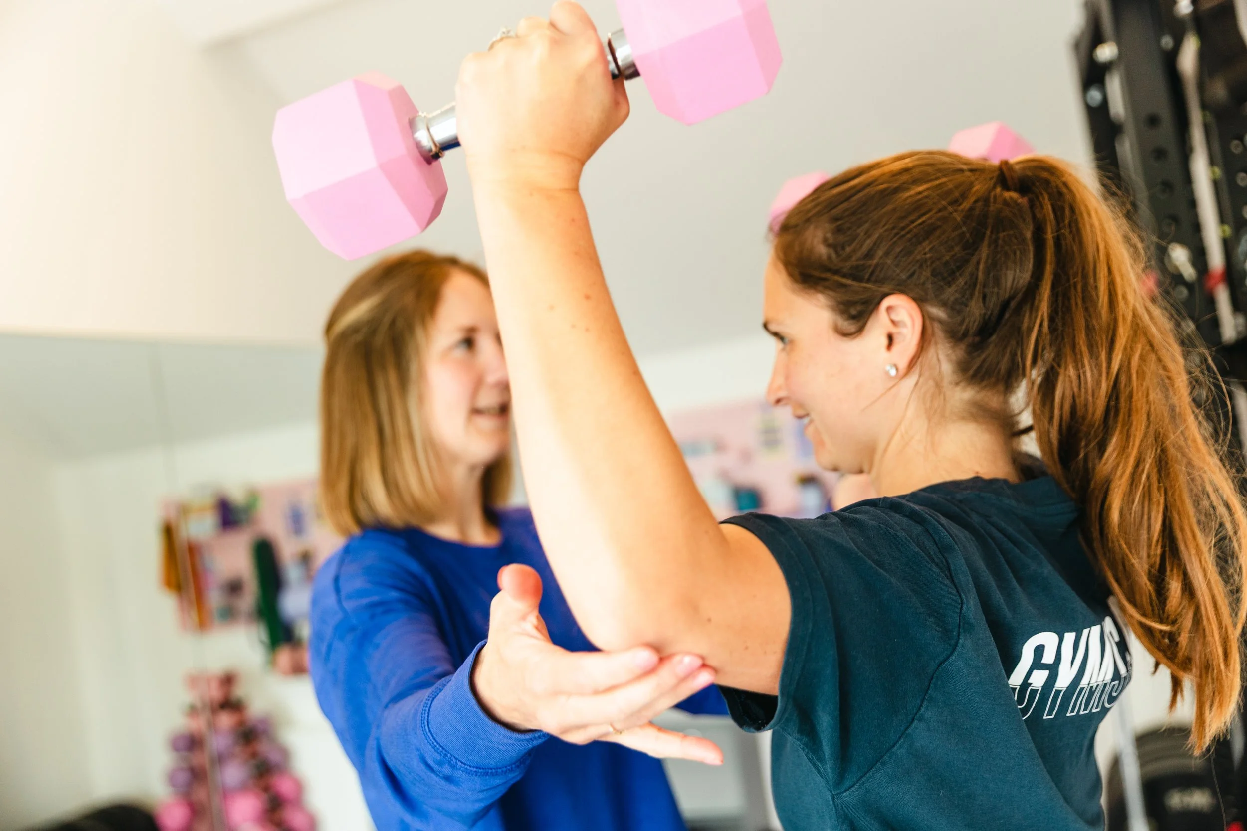 Two women are engaging in a workout, with one woman lifting pink dumbbells and the other spotting. They are smiling and appear to be in a fitness class or gym.