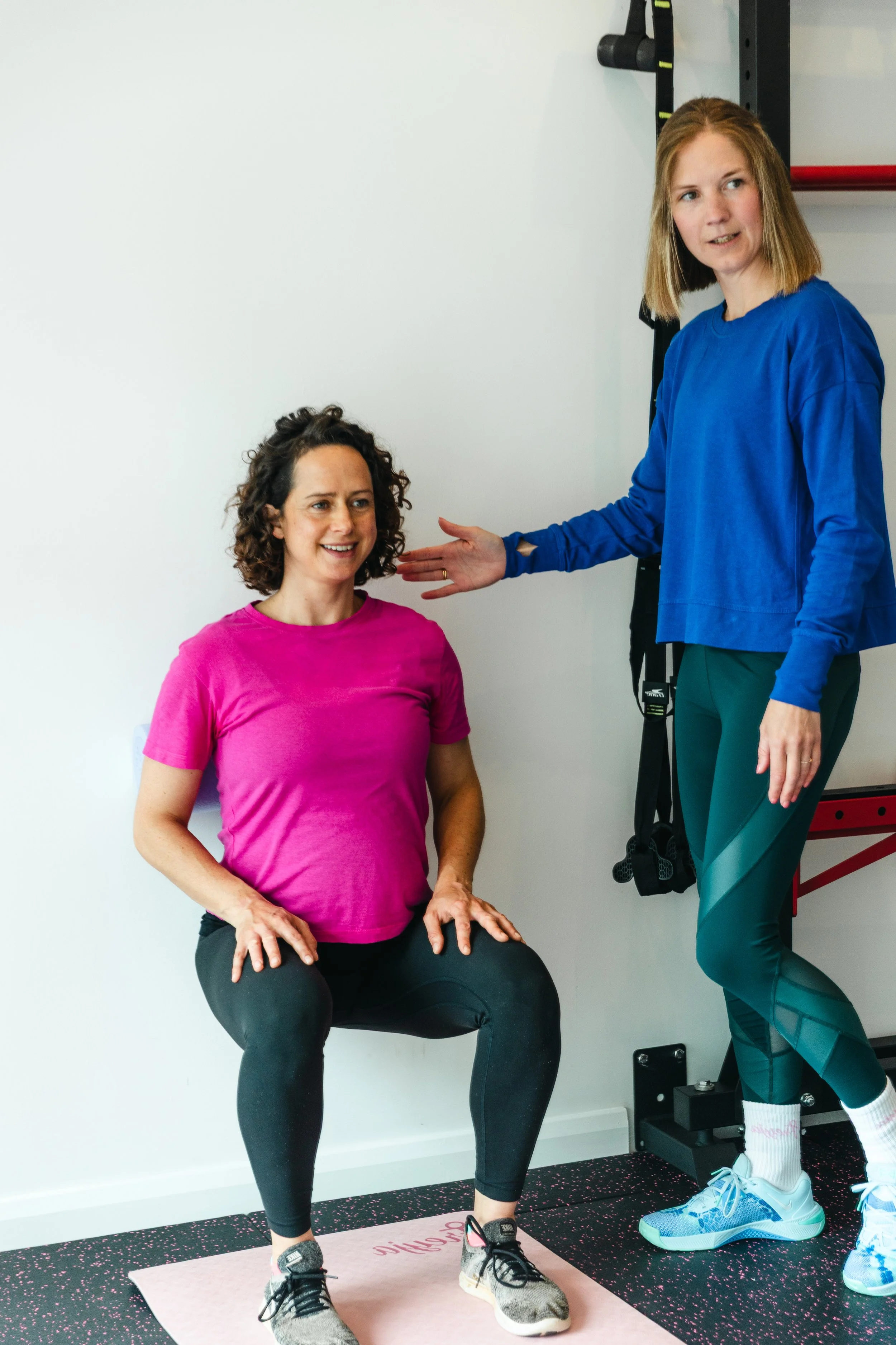 A woman in a pink t-shirt and black leggings sitting on an exercise ball, smiling, with a workout partner in a blue long-sleeve shirt and green leggings standing beside her, with a hand near her head, in a fitness studio with white walls and workout equipment.