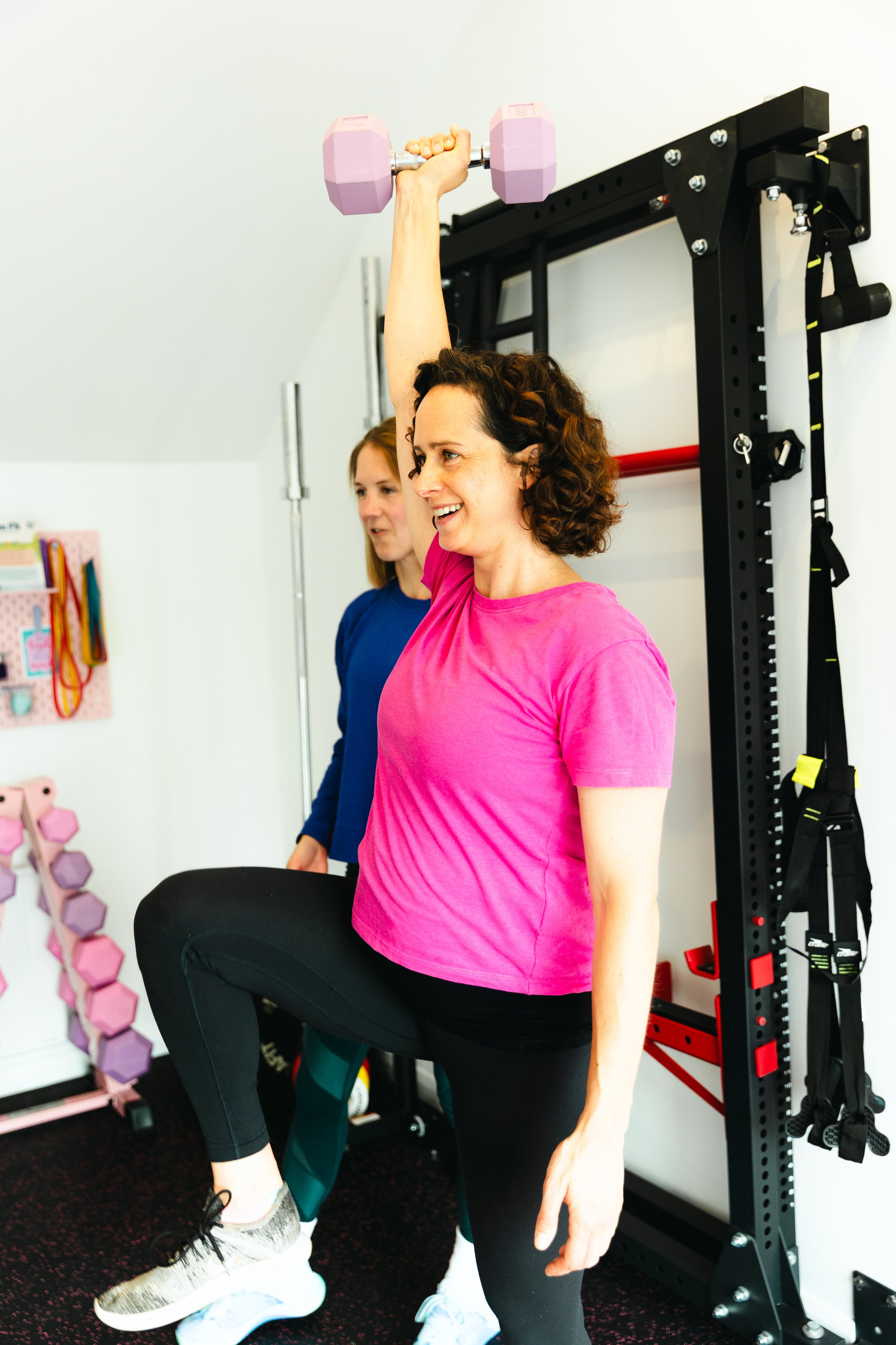 A trainer working with a pregnant lady in a gym, one woman lifting her knee and the other holding a pink dumbbell overhead.