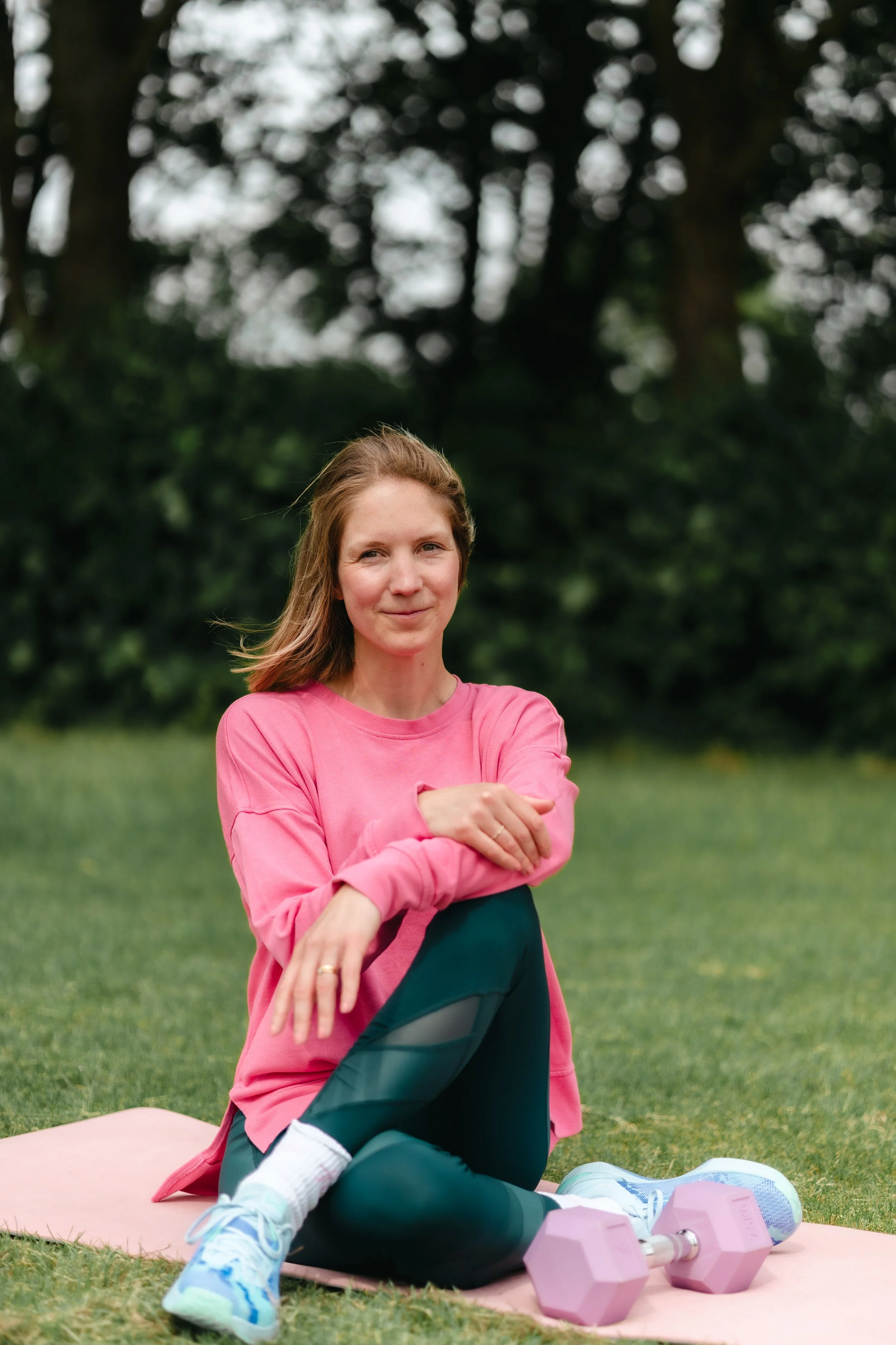 A woman in a pink shirt and black leggings sitting on a pink exercise mat outdoors, stretching with her arms crossed. There are pink dumbbells and white athletic shoes nearby on the grass, with a background of trees and greenery.