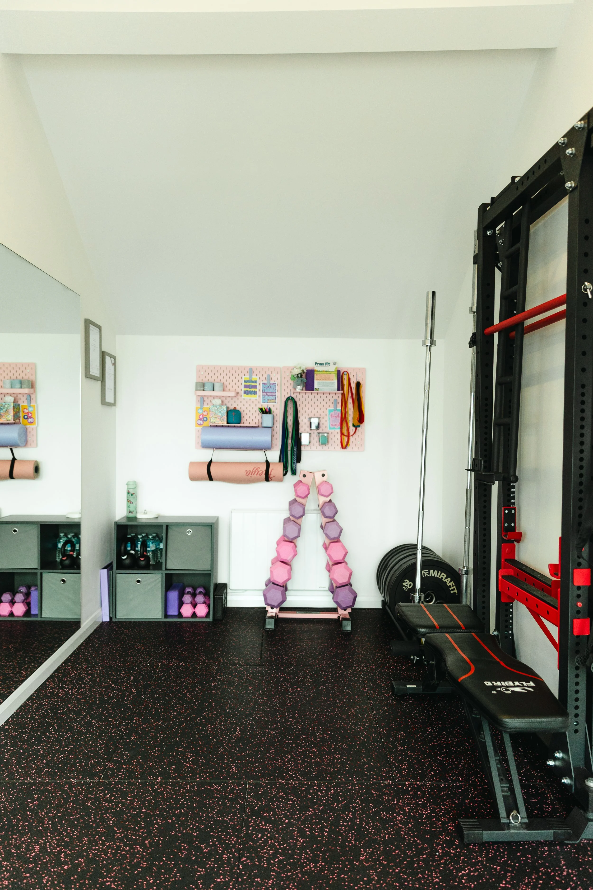 A home gym with pink and gray workout equipment. There are pink hexagonal dumbbells stacked on a rack, resistance bands hanging on wall hooks, and a pink patterned pegboard with small shelves and storage containers. The black rubber floor has pink speckles, and there are shelves with kettlebells and other fitness gear. A black weight plate and a black weight bench are also visible.
