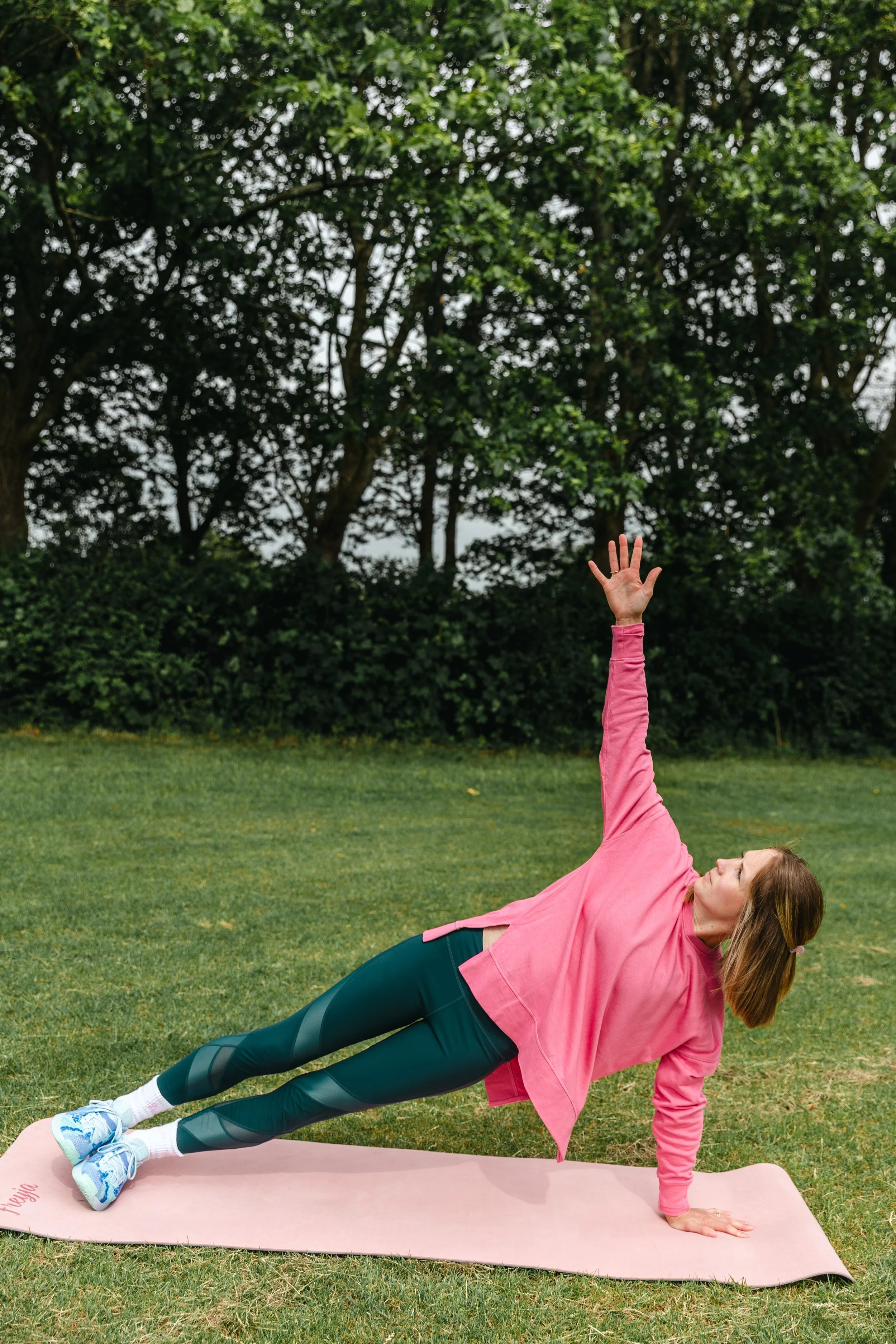 A woman doing a side plank exercise outdoors on a pink yoga mat, wearing a pink workout hoodie, black leggings with patterned panels, and sneakers, with a background of green trees and grass.