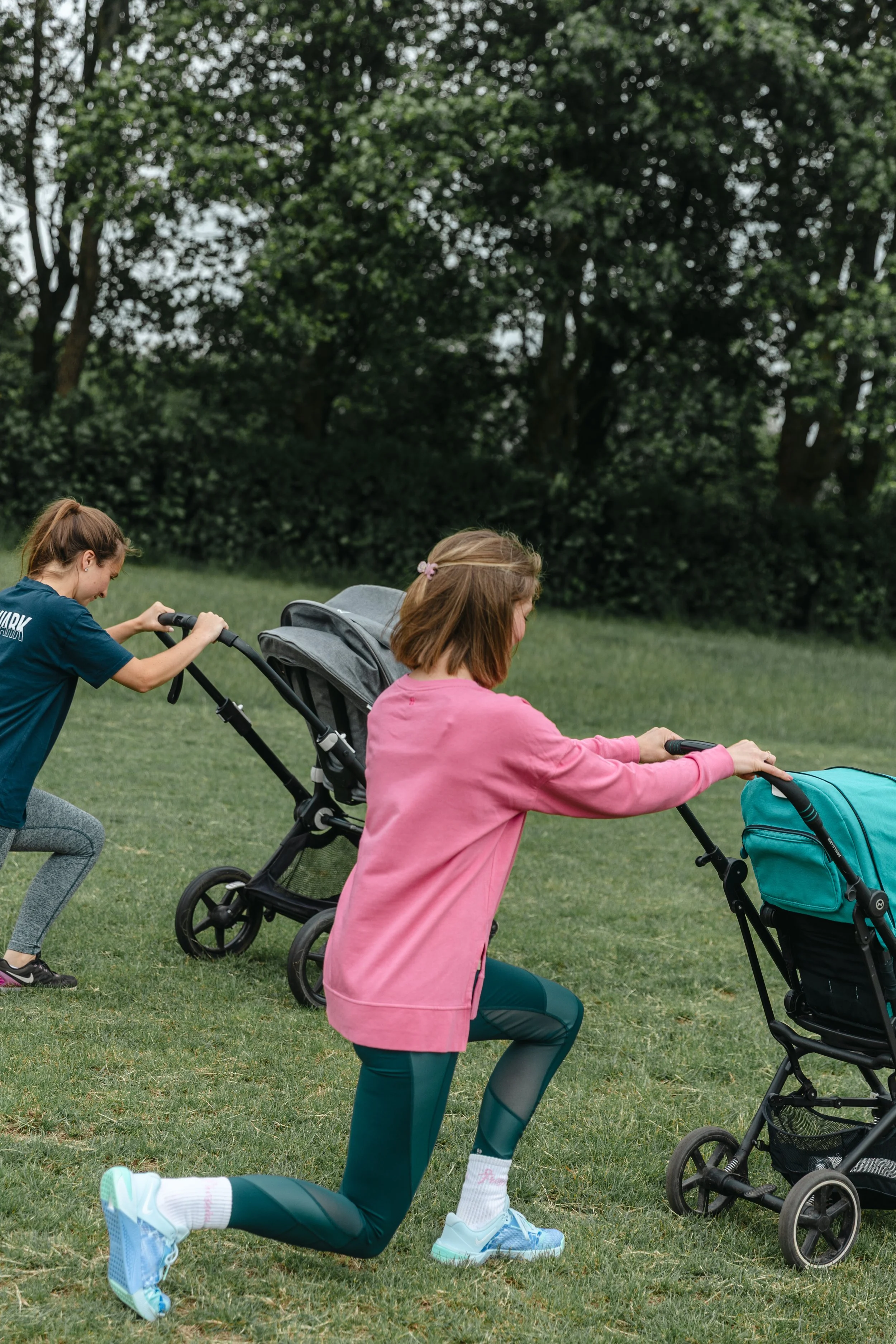 Two women are outside on a grassy area with trees in the background. One woman, wearing a pink top and teal leggings, is kneeling on one knee and pushing a stroller. The other woman, wearing a blue top, is pushing a stroller and smiling.