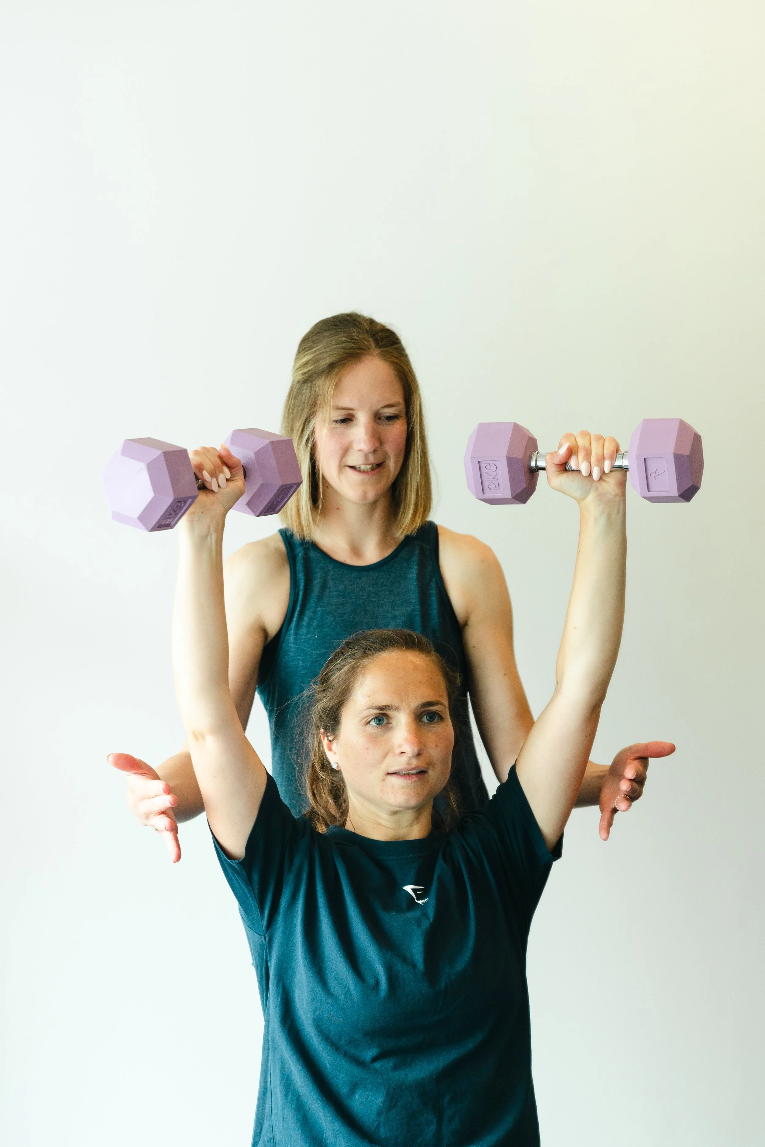 Two women perform a yoga pose with one woman standing behind the other, both holding light purple dumbbells, against a plain white background.