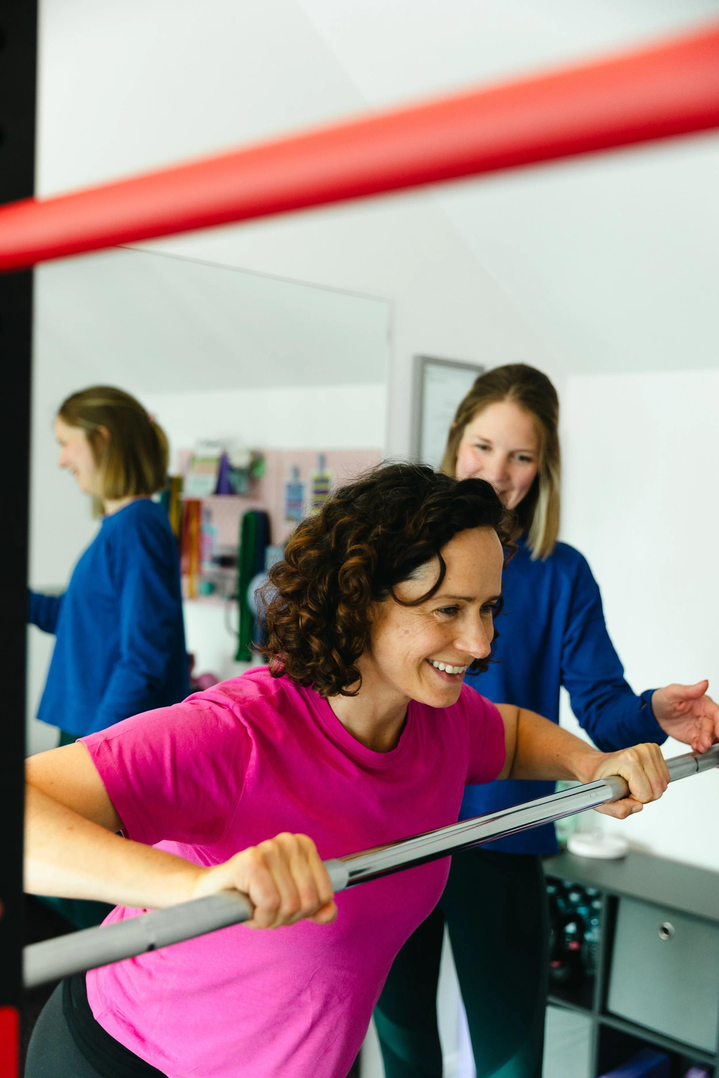 Women in a gym using horizontal bars for exercise, wearing workout clothes, with a woman in a pink shirt in the foreground and other women in blue shirts in the background.