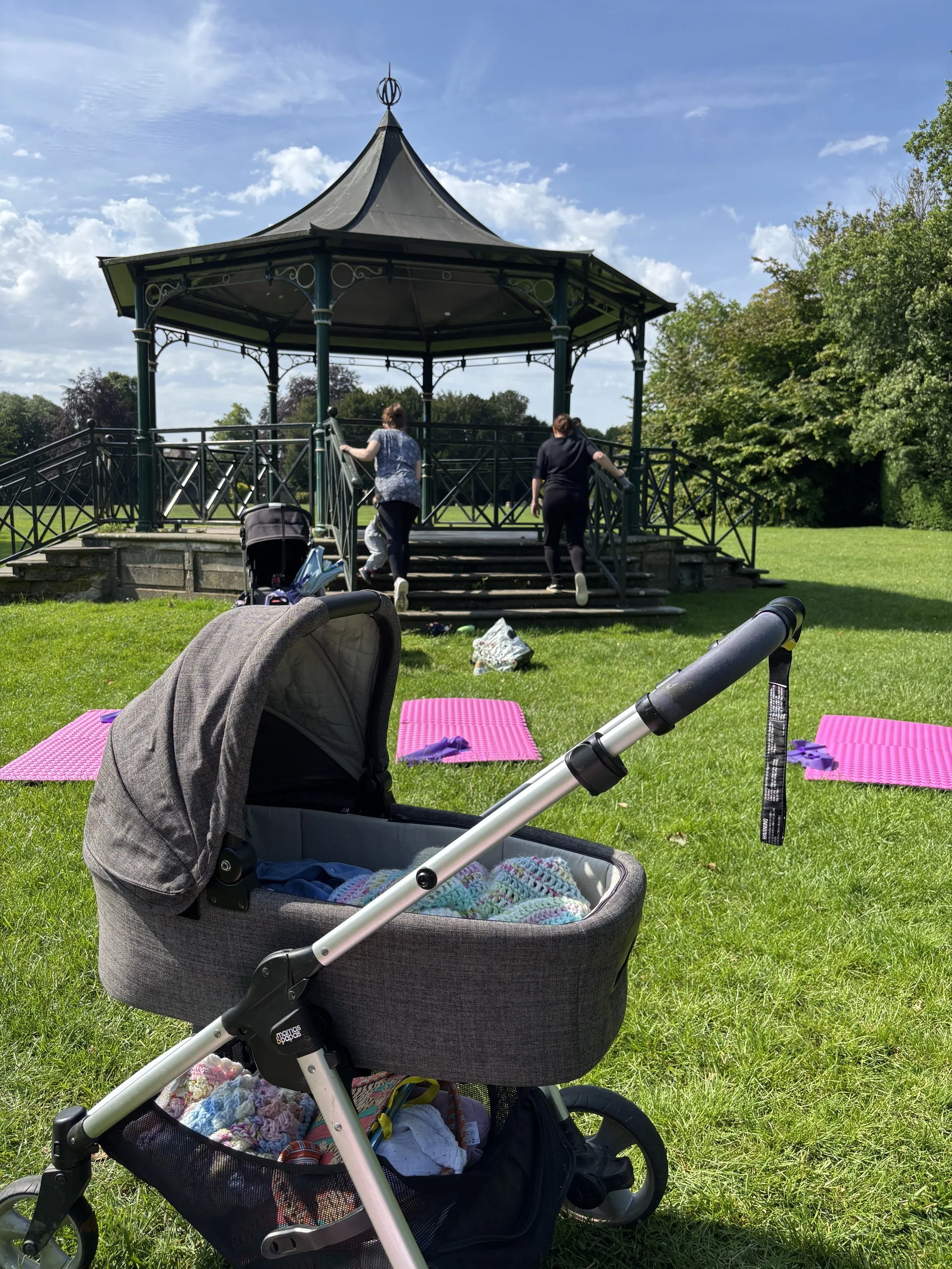 A park gazebo with people climbing the stairs, on green grass with pink yoga mats and baby strollers in the foreground, blue sky with clouds in the background.