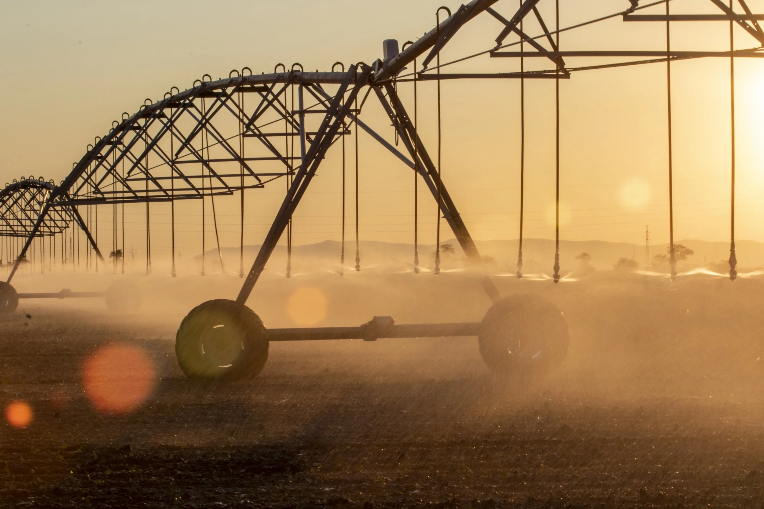 Large irrigation sprinklers watering a field at sunset with a warm orange glow.