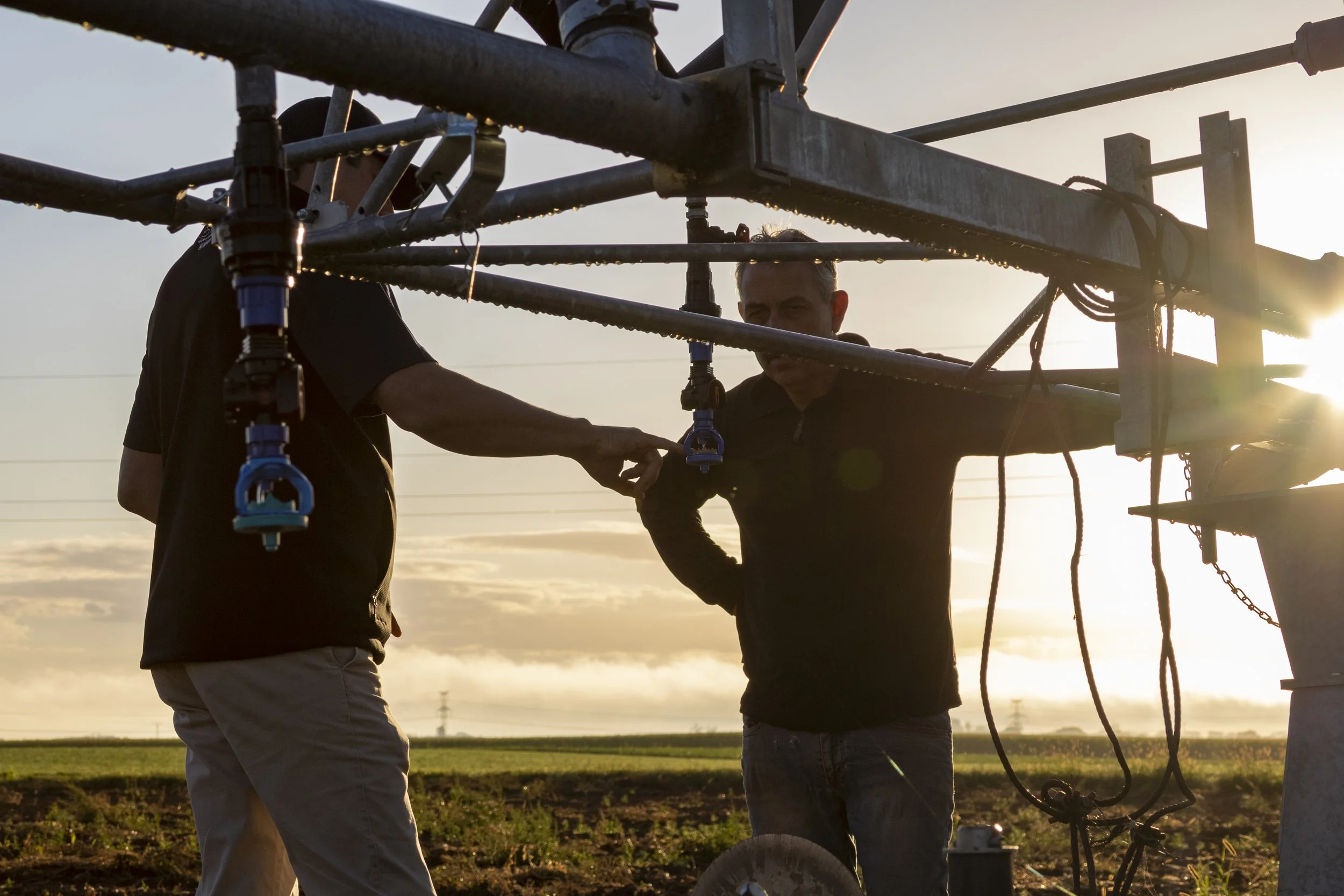 Two men working on a large mechanical structure outdoors during sunset or sunrise, with a field in the background.