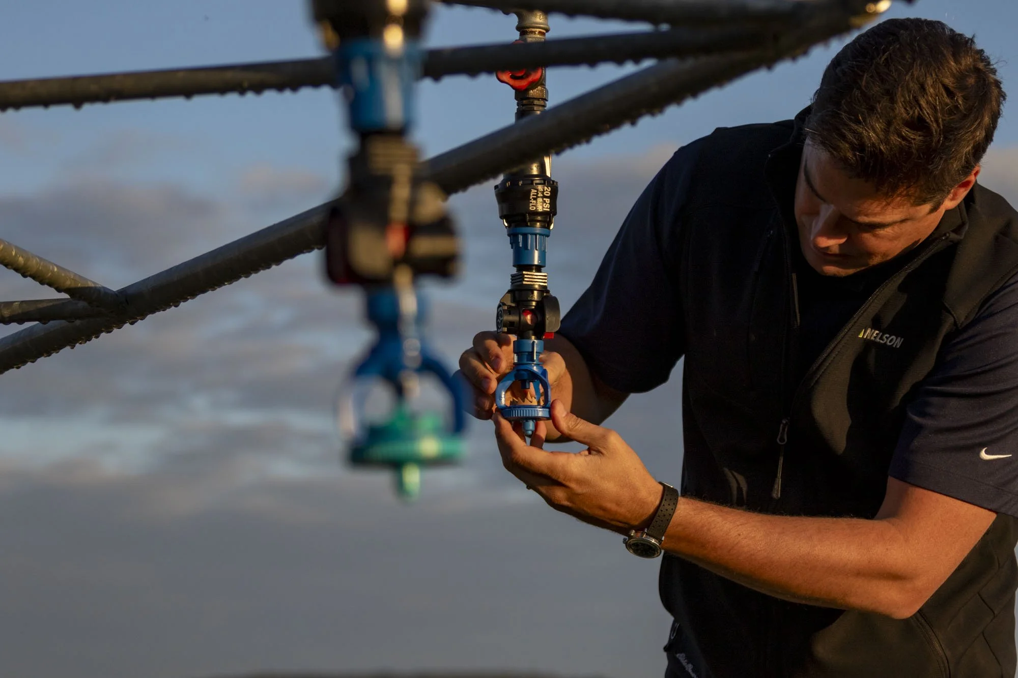 A man working on a mechanical assembly or device outdoors, tightening or adjusting a component, with parts of a metal structure and a cloudy sky in the background.
