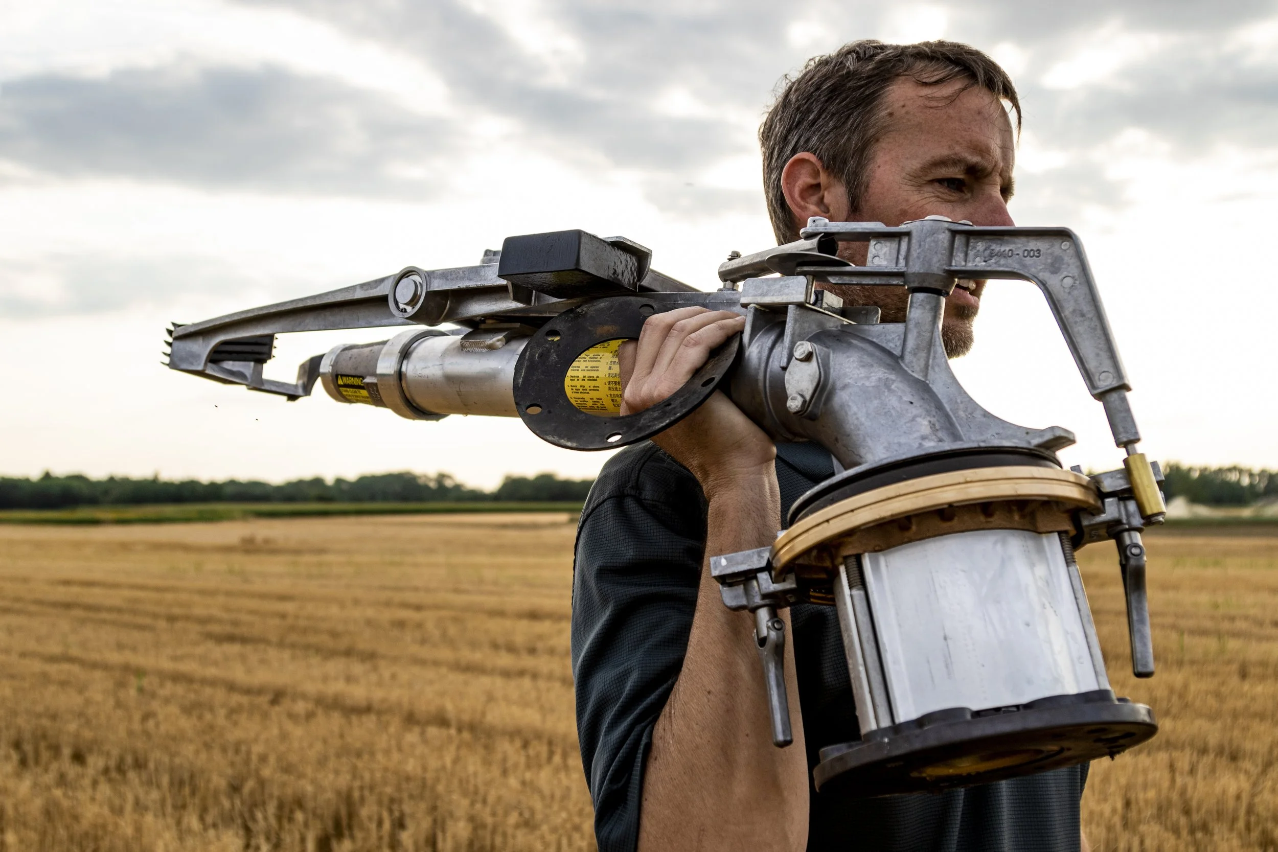 A man standing in a field carrying a large mechanical device on his shoulder.