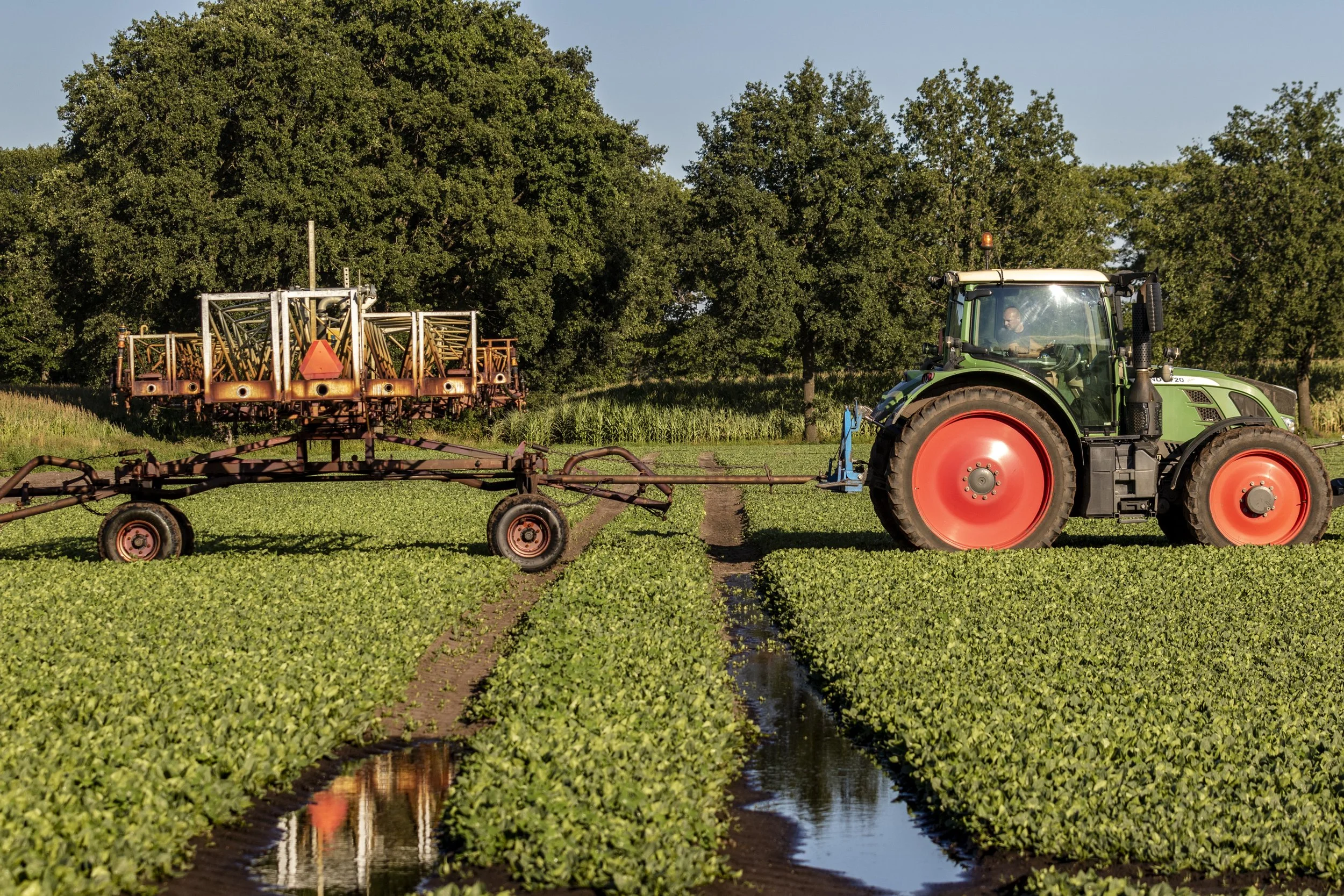 A tractor pulling a sprayer across a green field with water channels, surrounded by trees under a clear sky.