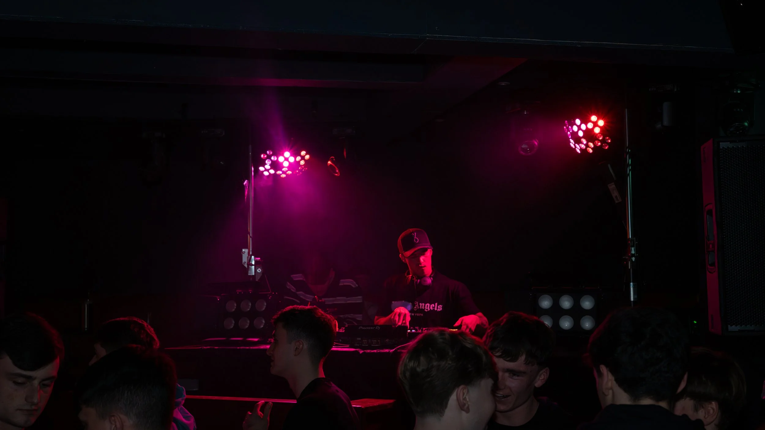 A DJ in black clothing and a black cap stands behind a mixer in a dark club with purple and pink stage lighting, while a crowd of young people, mostly male, dance and socialize in front.