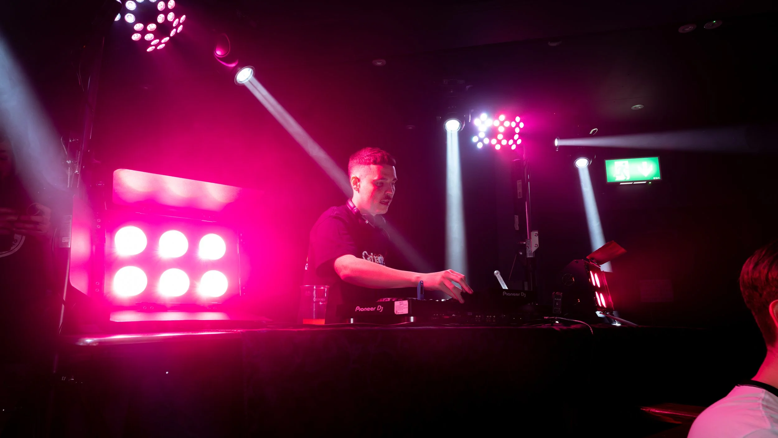 A DJ performing at a nightclub with pink and white lights, wearing headphones and a T-shirt, standing behind a Pioneer DJ controller, with a beer glass on the table.
