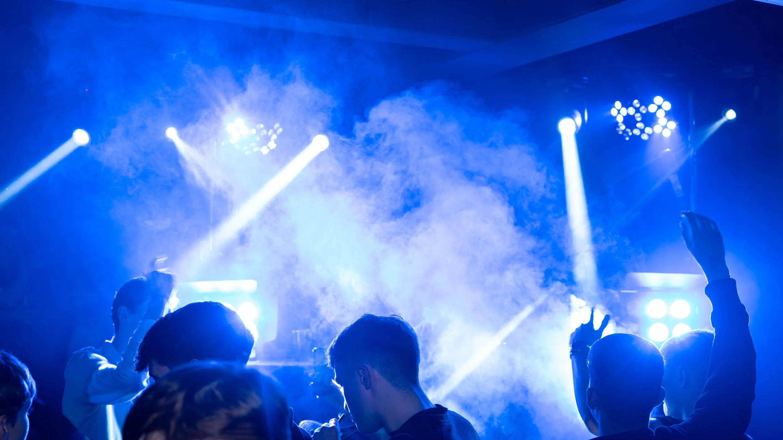 Crowd of people at a concert or nightclub with colorful stage lighting and fog, raising their hands and enjoying the music.