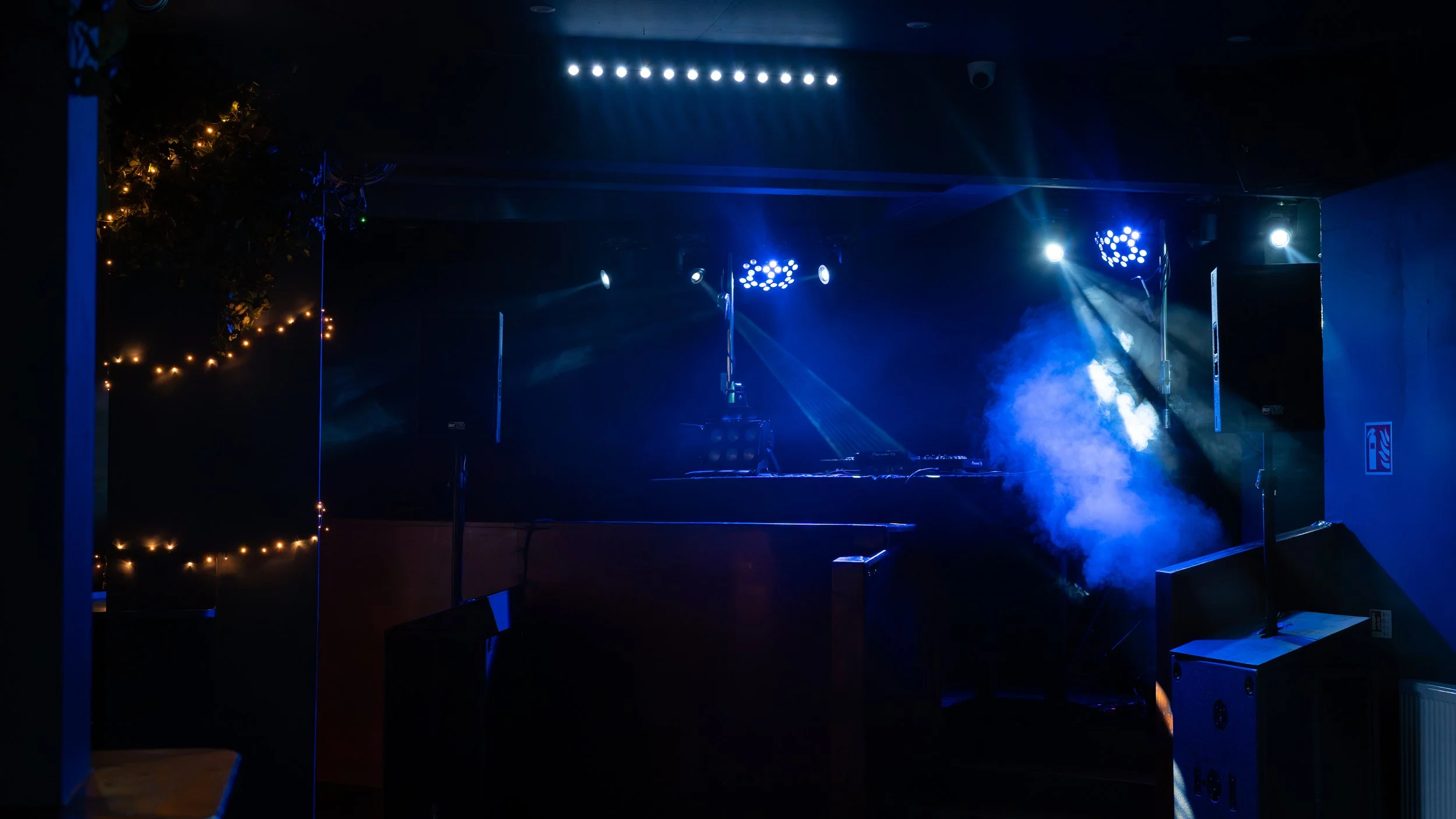 Empty stage with DJ equipment, blue and white stage lights, and fog effects, set up for a nightclub or concert event.
