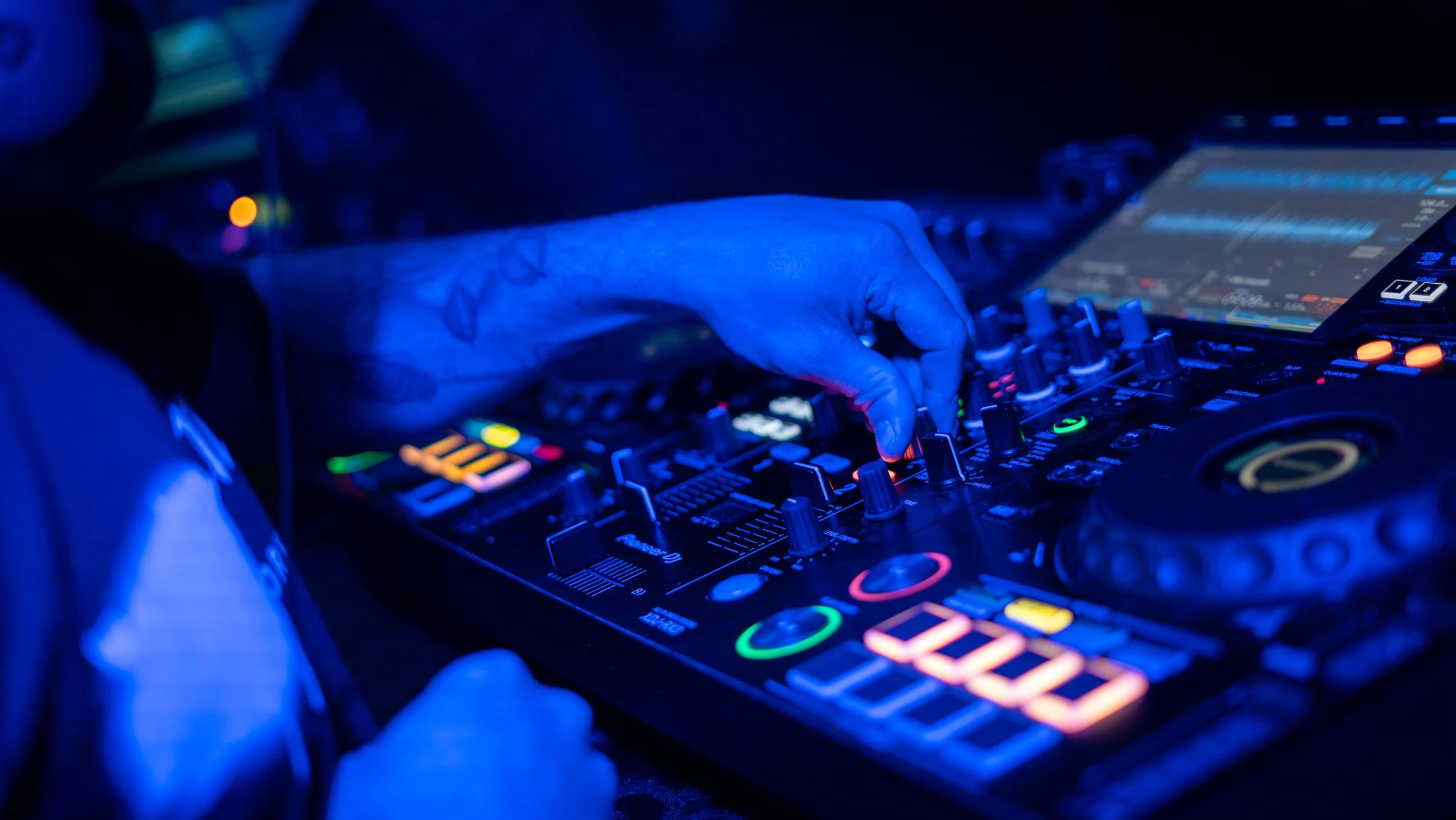 A DJ's hand adjusting controls on a professional DJ mixer and controller with colorful illuminated buttons, in a dark setting.