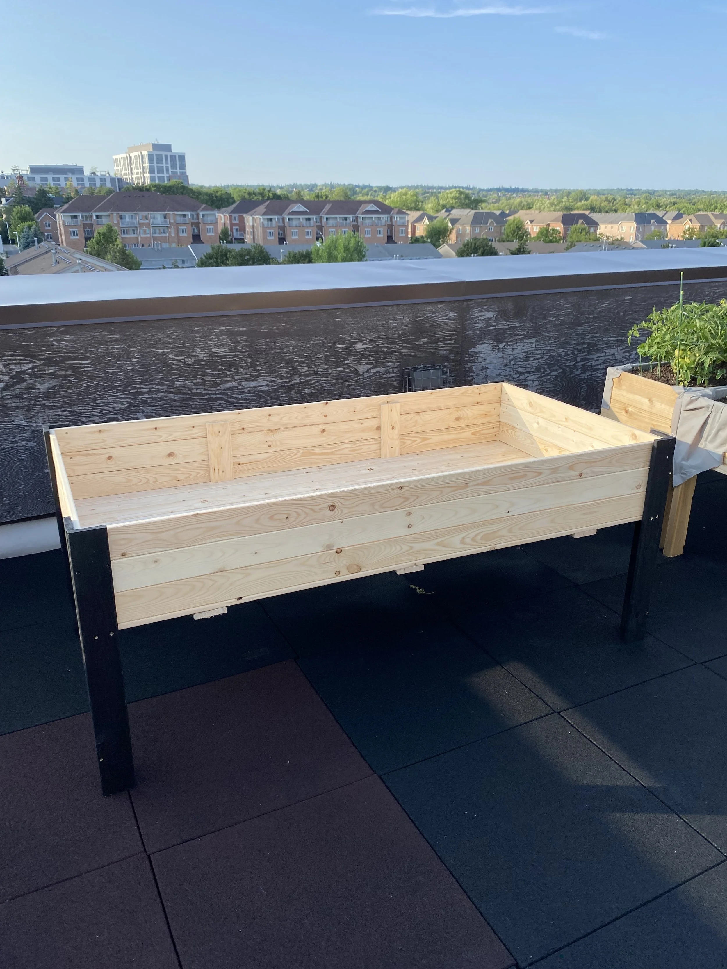 A wooden custom planter box on a rooftop balcony with city apartment buildings and trees in the background under a clear sky.