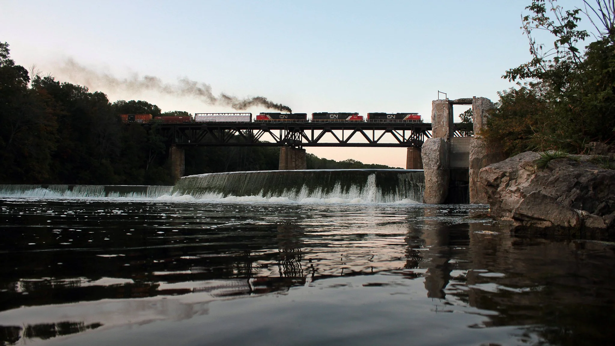 A train crossing a bridge over a waterfall on a river, with trees and rocks on the banks, and smoke coming from the train engine.